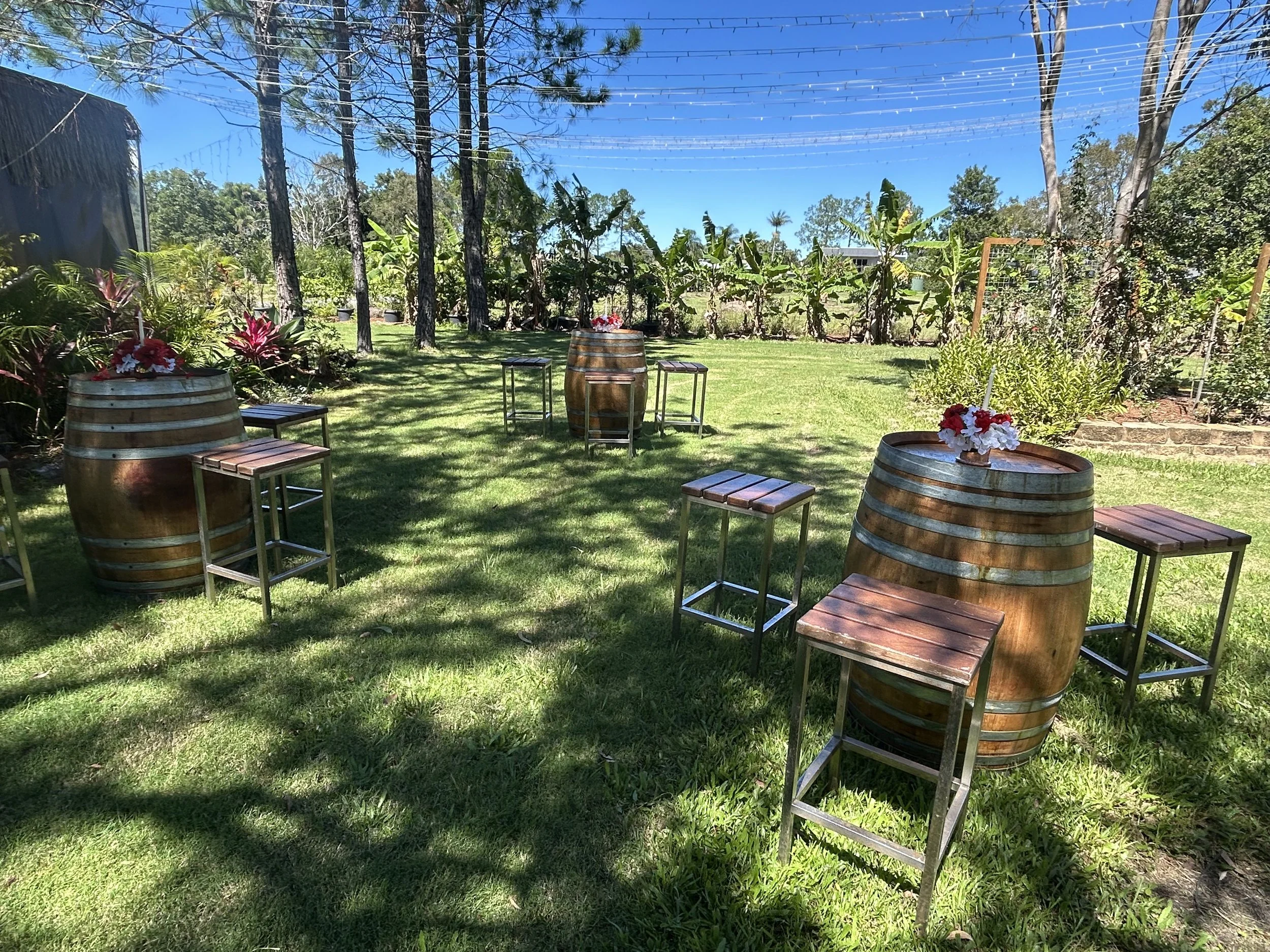Outdoor garden setting with wine barrels used as tables, topped with flower arrangements, surrounded by wooden stools, with trees and lush greenery in the background under a clear blue sky.
