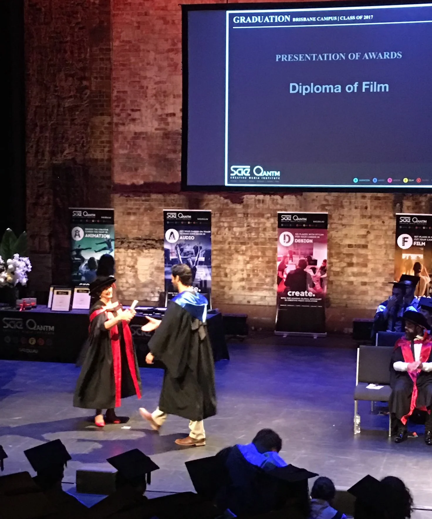 Graduation ceremony on stage with graduates in academic robes. One graduate in a black robe with red accents is receiving a diploma from another graduate in a black robe with a blue hood. There are seated academic officials and banners in the background. A large screen displays text about the graduation event.