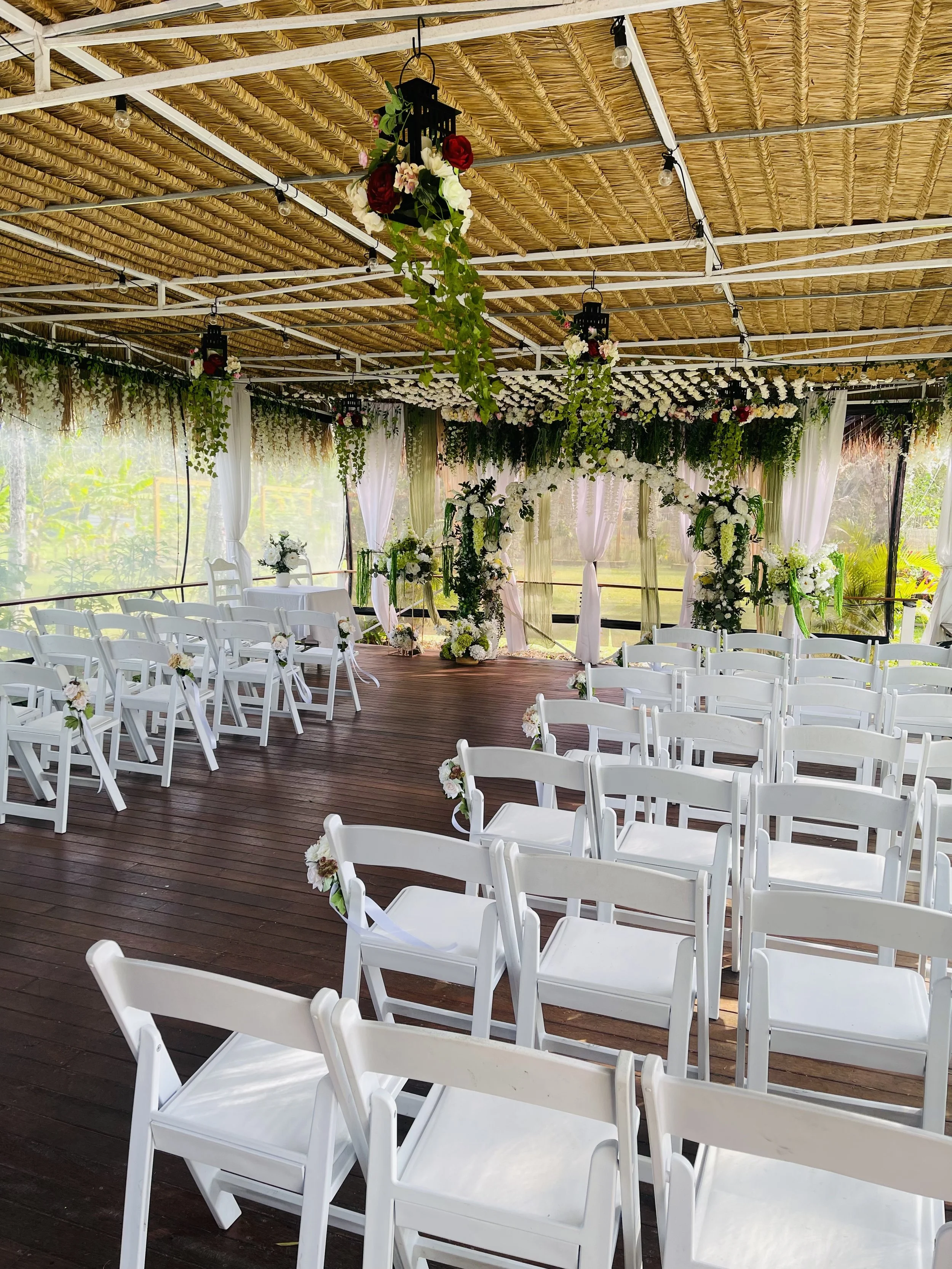 Indoor wedding venue decorated with white chairs, floral arrangements, and hanging lanterns with flowers and greenery. The ceiling has a woven thatch covering and string lights.