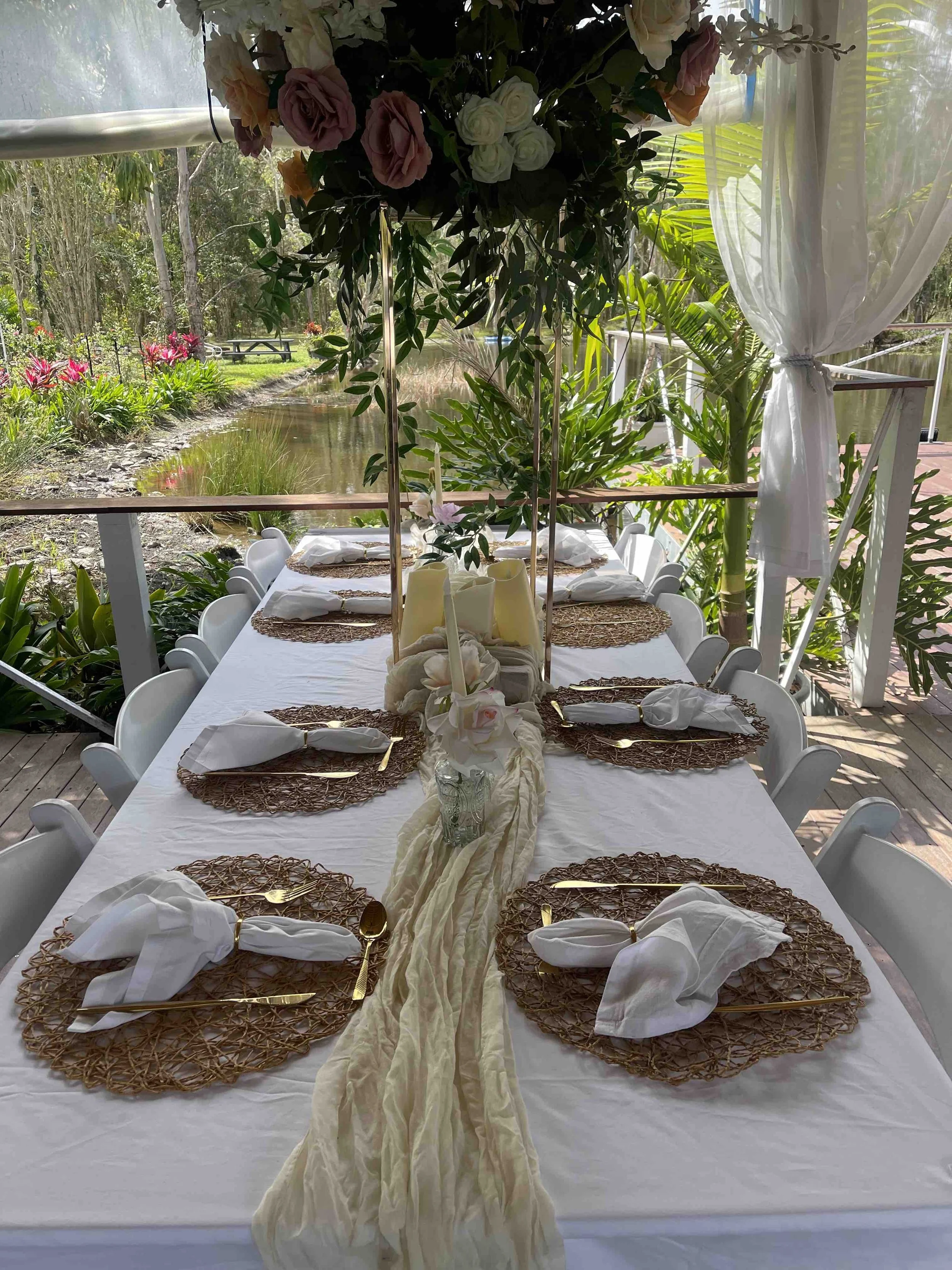 Outdoor table set for a celebration, with a white tablecloth, woven placemats, gold cutlery, and napkins folded into a decorative shape. The table is decorated with a floral centerpiece and surrounded by white chairs. The background features lush gre