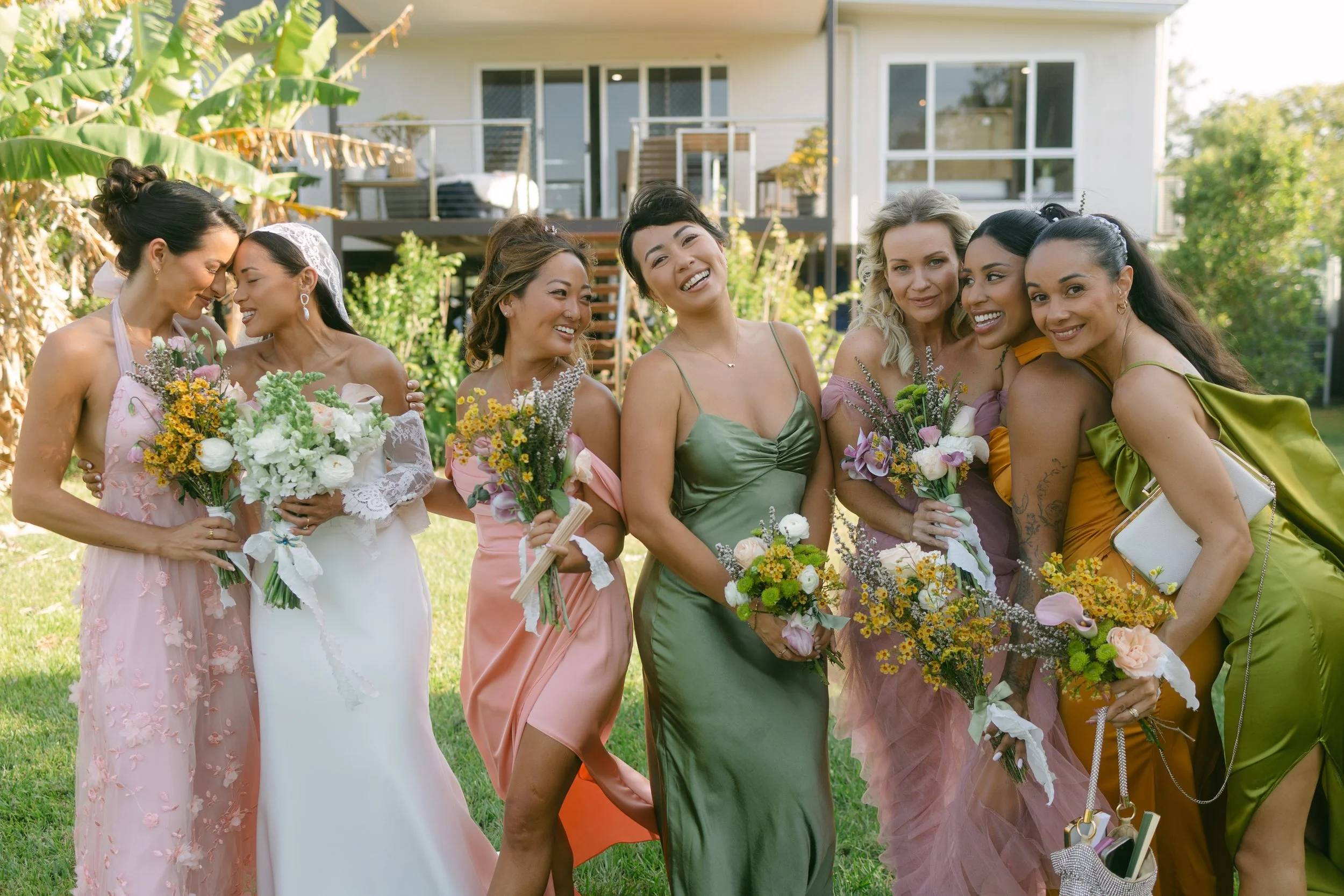 Group of women dressed in colorful dresses, holding flowers, gathered outdoors in a garden with house in the background.