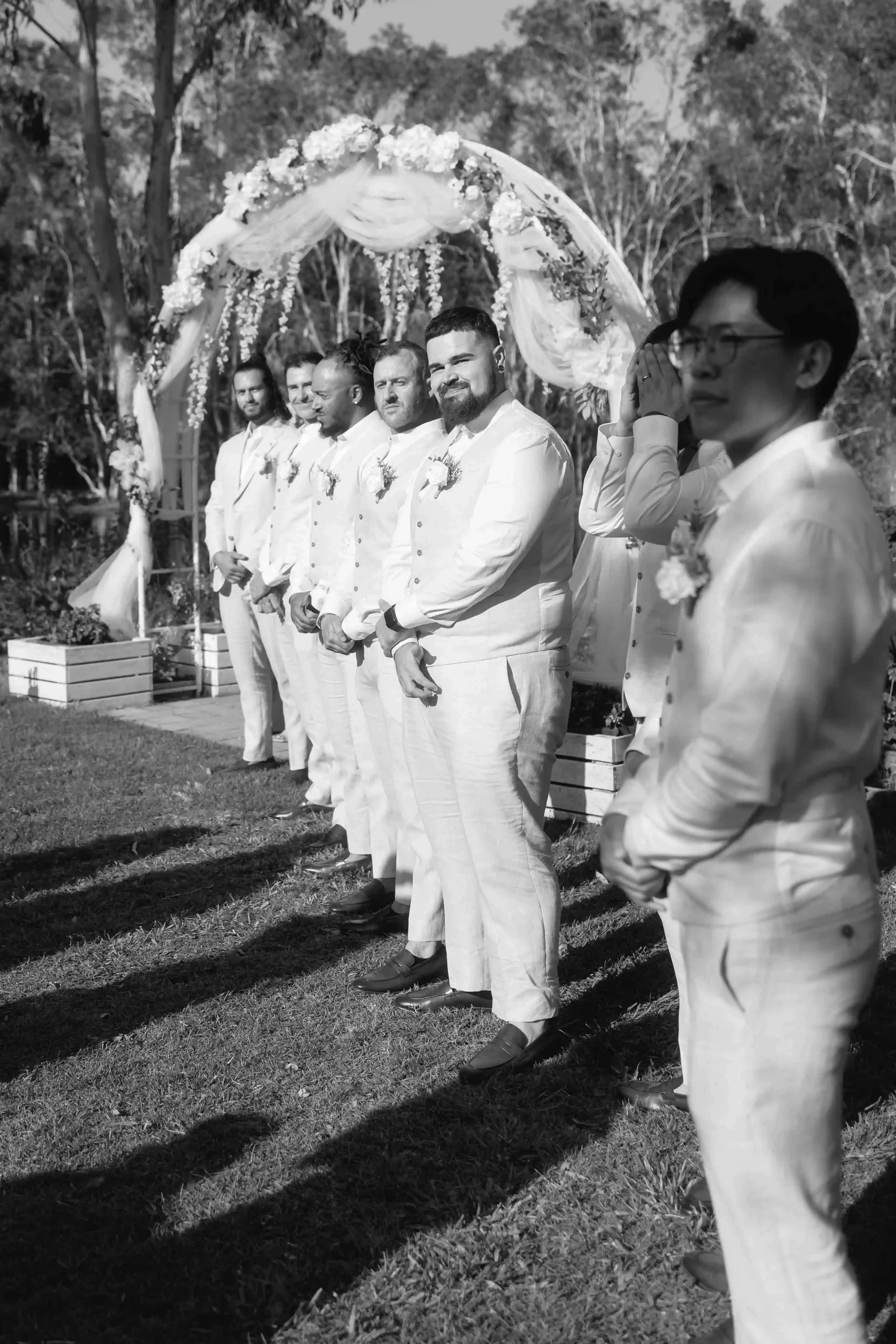 A black and white photo of a wedding ceremony outdoors, with seven men dressed in white suits, standing in a row under a floral arch, some with hands clasped or covering their faces, with trees in the background.