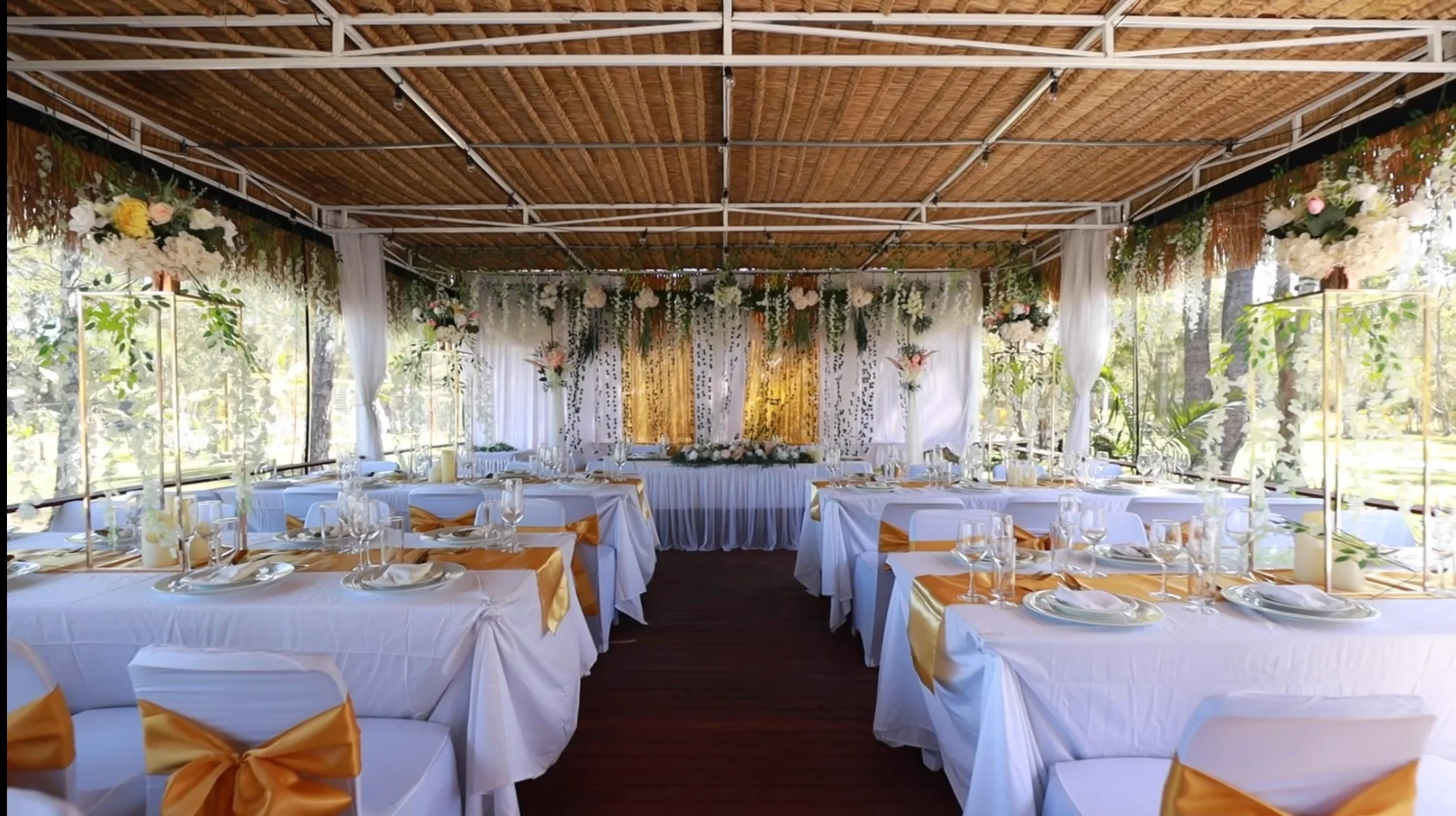 Elegant outdoor wedding reception setup with white tables, gold accents, floral centerpieces, and hanging flowers, under a thatched ceiling and surrounded by trees.