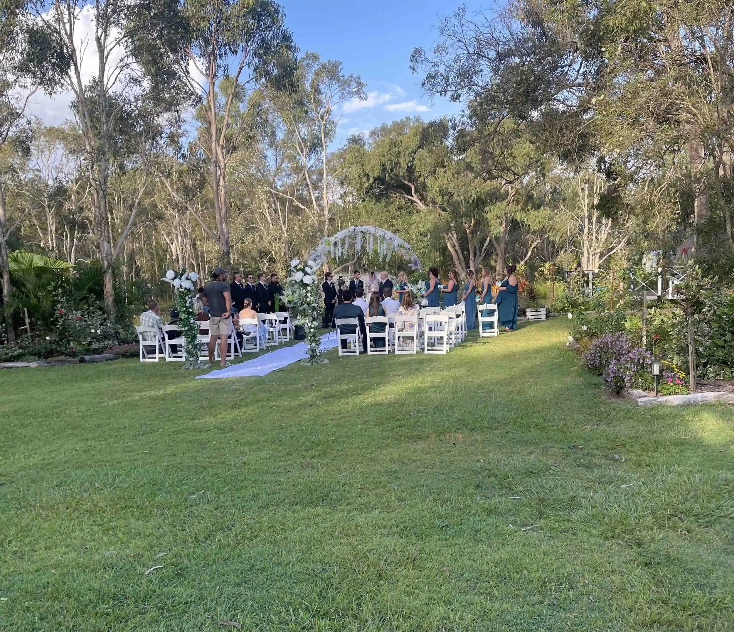 Outdoor wedding ceremony setup in a garden with white chairs arranged in rows, floral decorations, and a floral arch, surrounded by trees and greenery under a clear blue sky.
