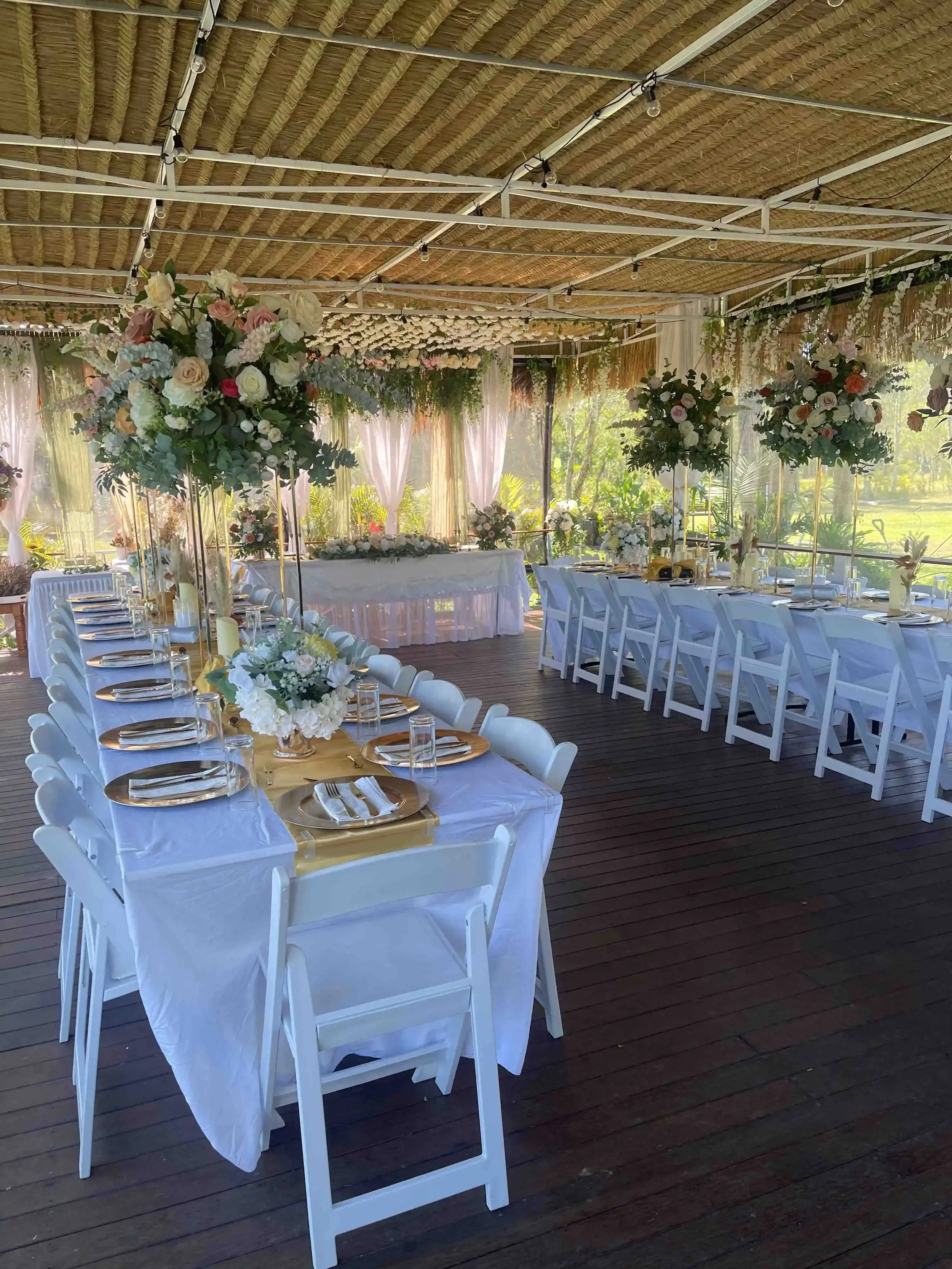 Wedding reception setup with long tables covered in white tablecloths, decorated with floral centerpieces, and set with plates, glasses, and cutlery. The venue features a wooden ceiling and large windows with pink curtains, overlooking greenery outsi