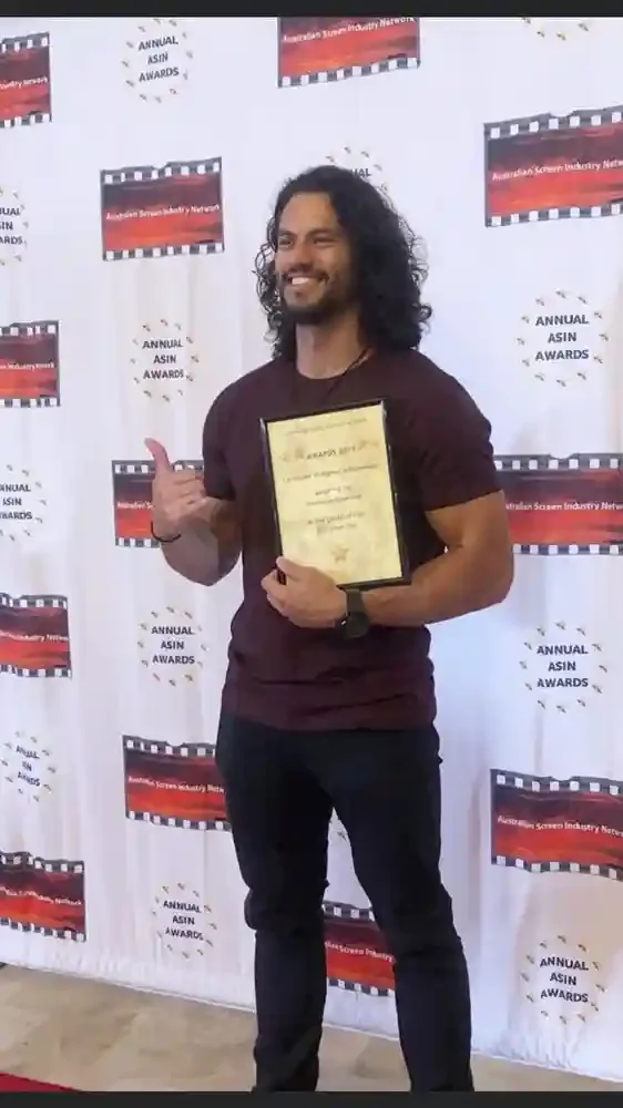 Man with long curly hair and beard holding a framed award, smiling, attending the Annual ASIN Awards in front of a backdrop with logos.