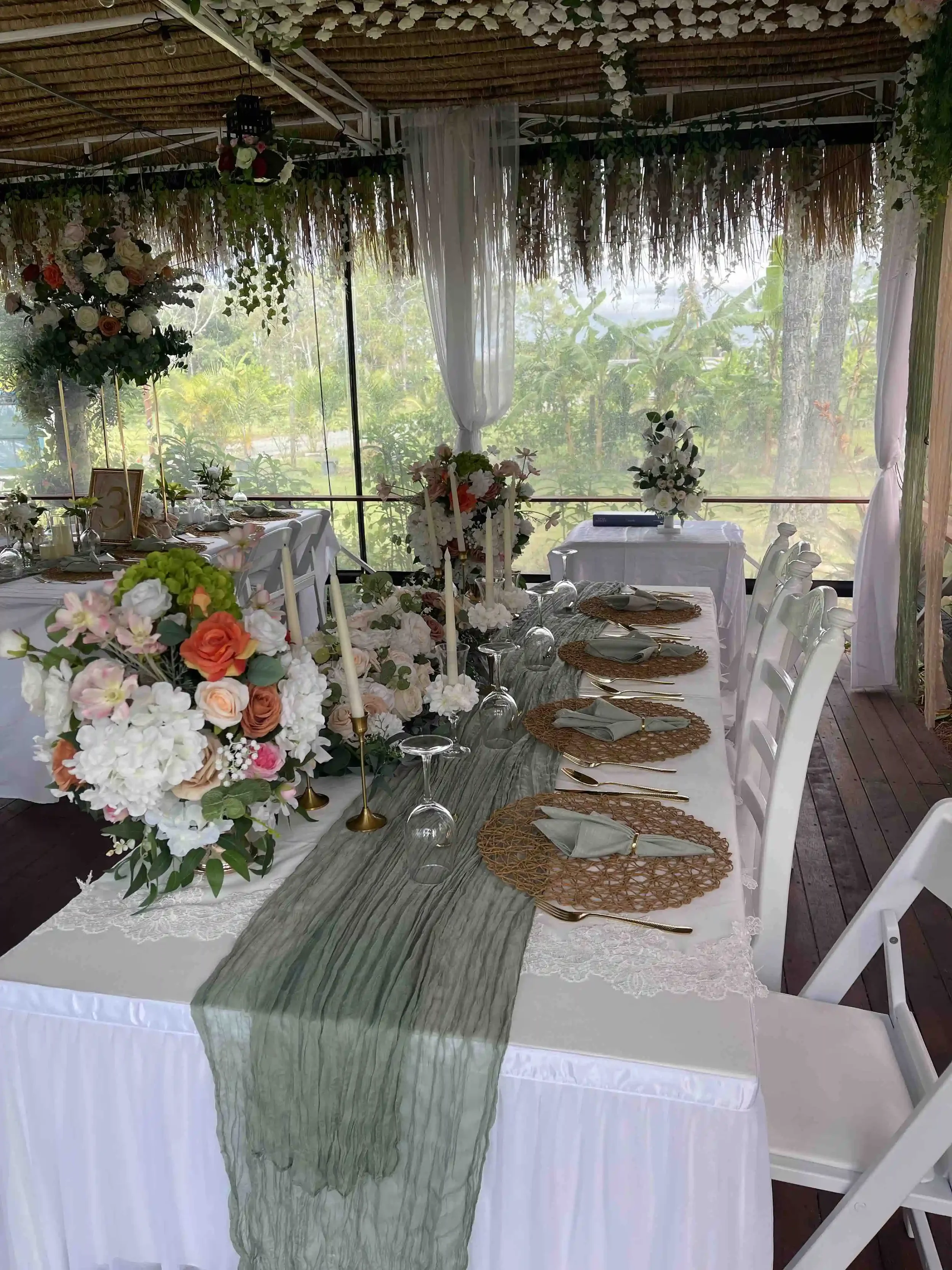Decorated dining table set for a celebration, featuring floral centerpieces, candles, woven placemats, and elegant tableware, with lush greenery and white curtains in the background.