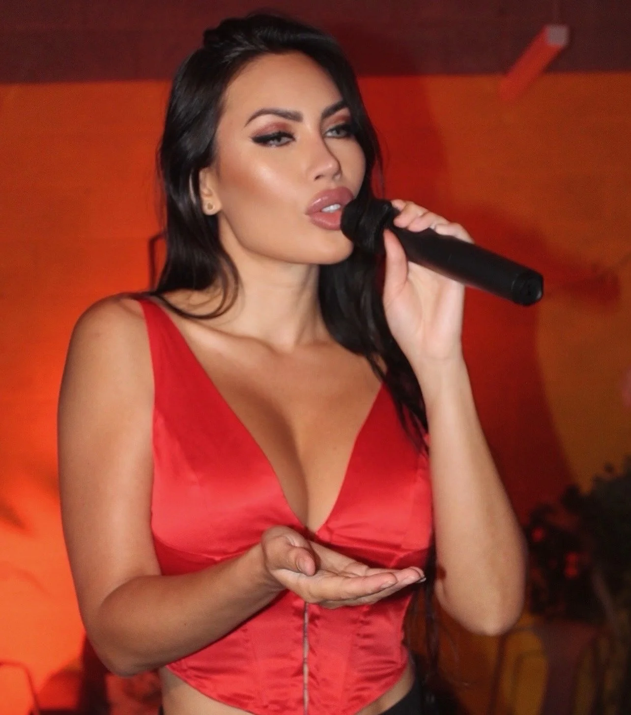 Woman with dark hair wearing a red top, holding a microphone.