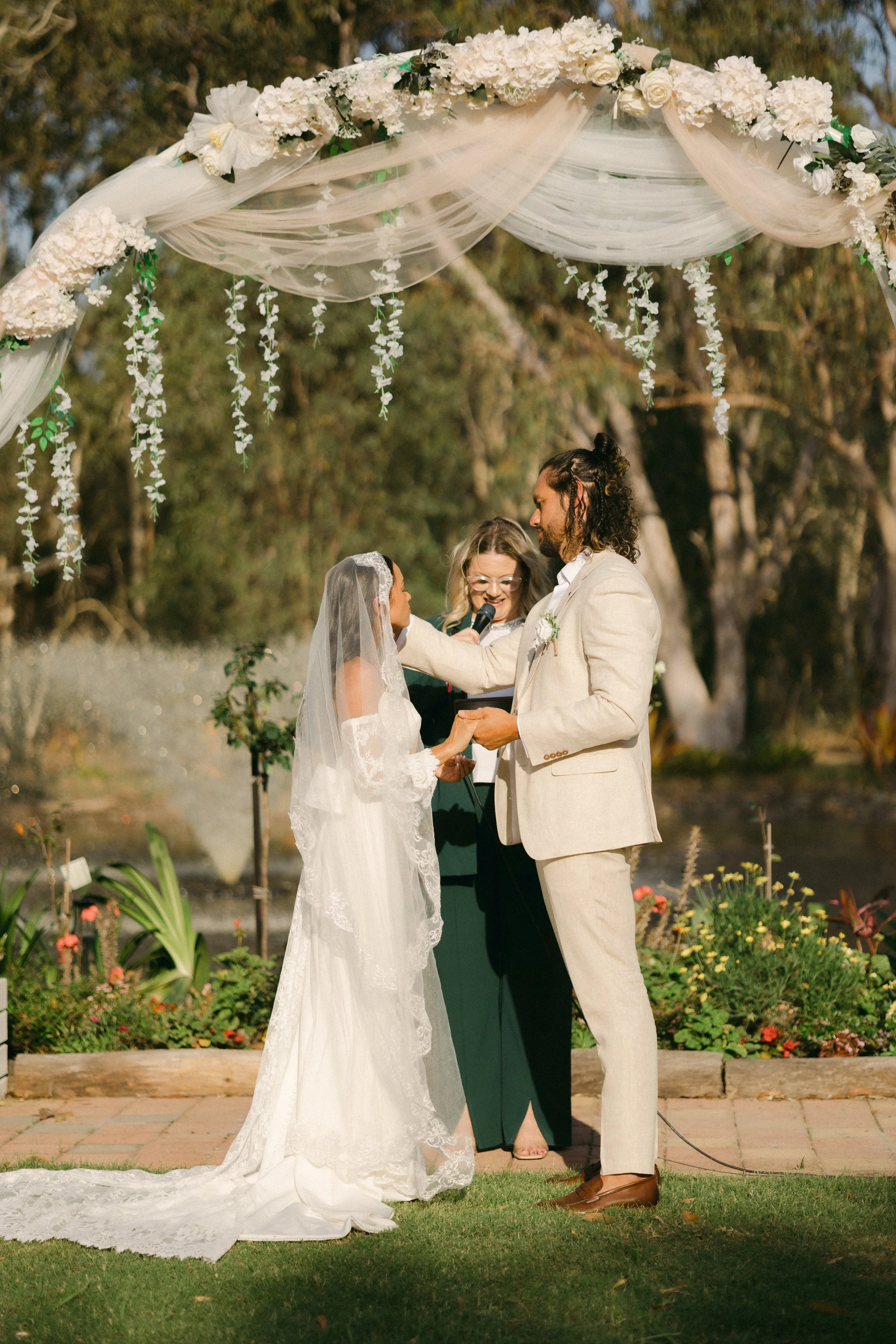 A couple getting married outdoors under a floral arch with an officiant, with a garden and trees in the background.