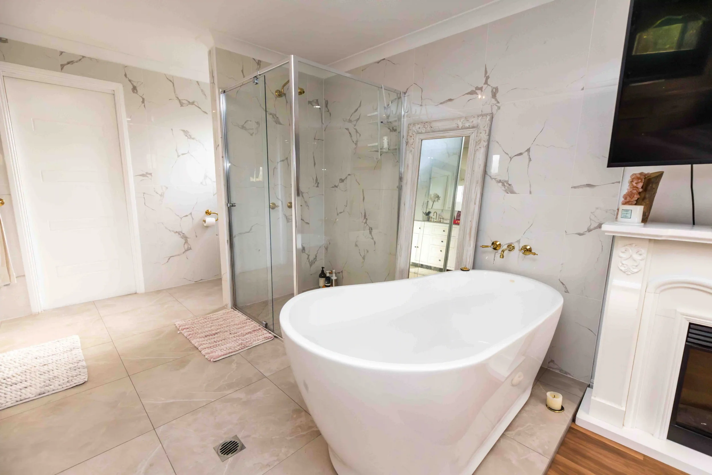 A modern bathroom with a freestanding bathtub, marble wall tiles, a glass shower enclosure, a large mirror, and white cabinetry. There are gold fixtures and a pink rug on the light-colored tiled floor.