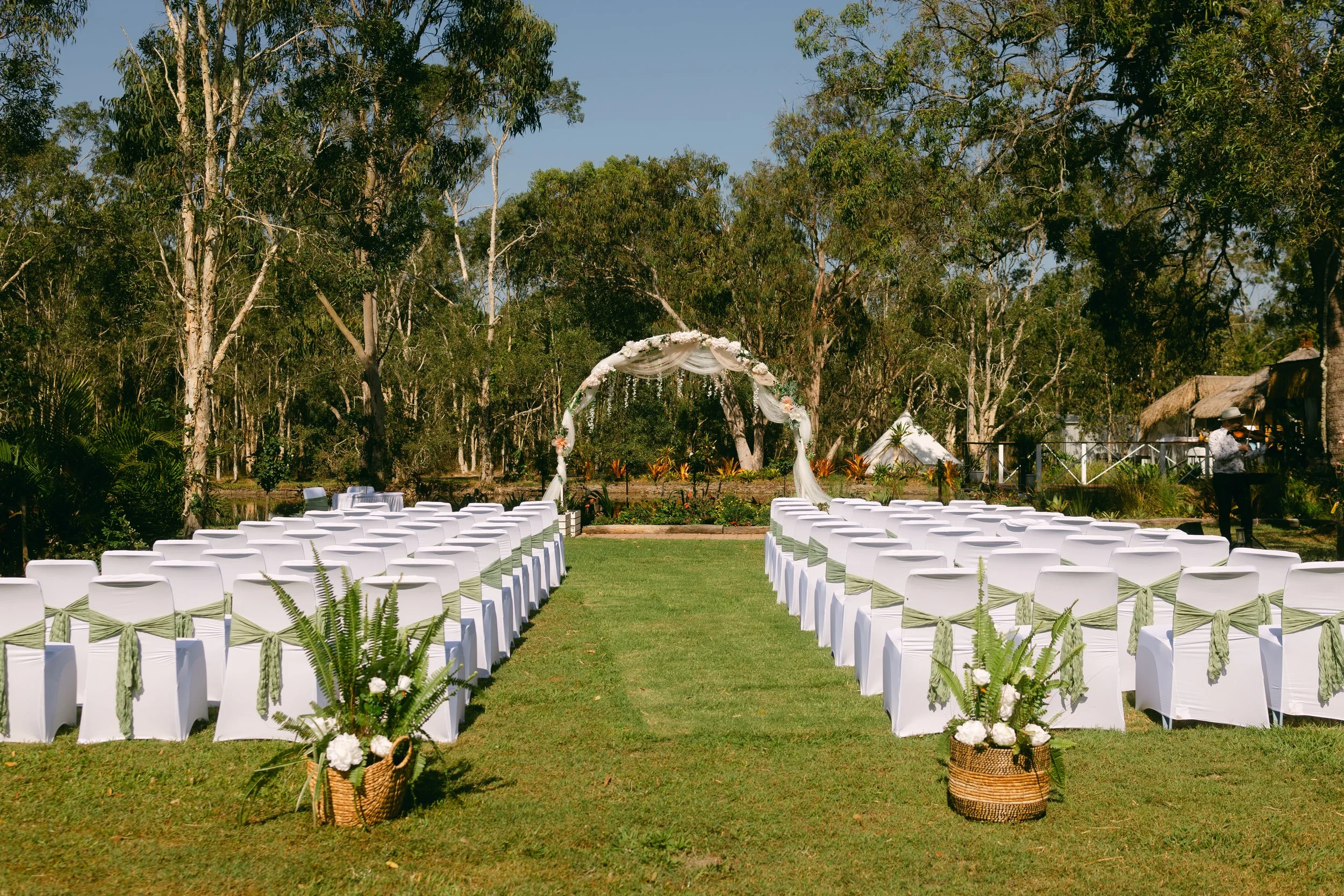Outdoor wedding ceremony setup with white chairs decorated with green sashes, an arch with white fabric and flowers, and potted plants on a grassy area
