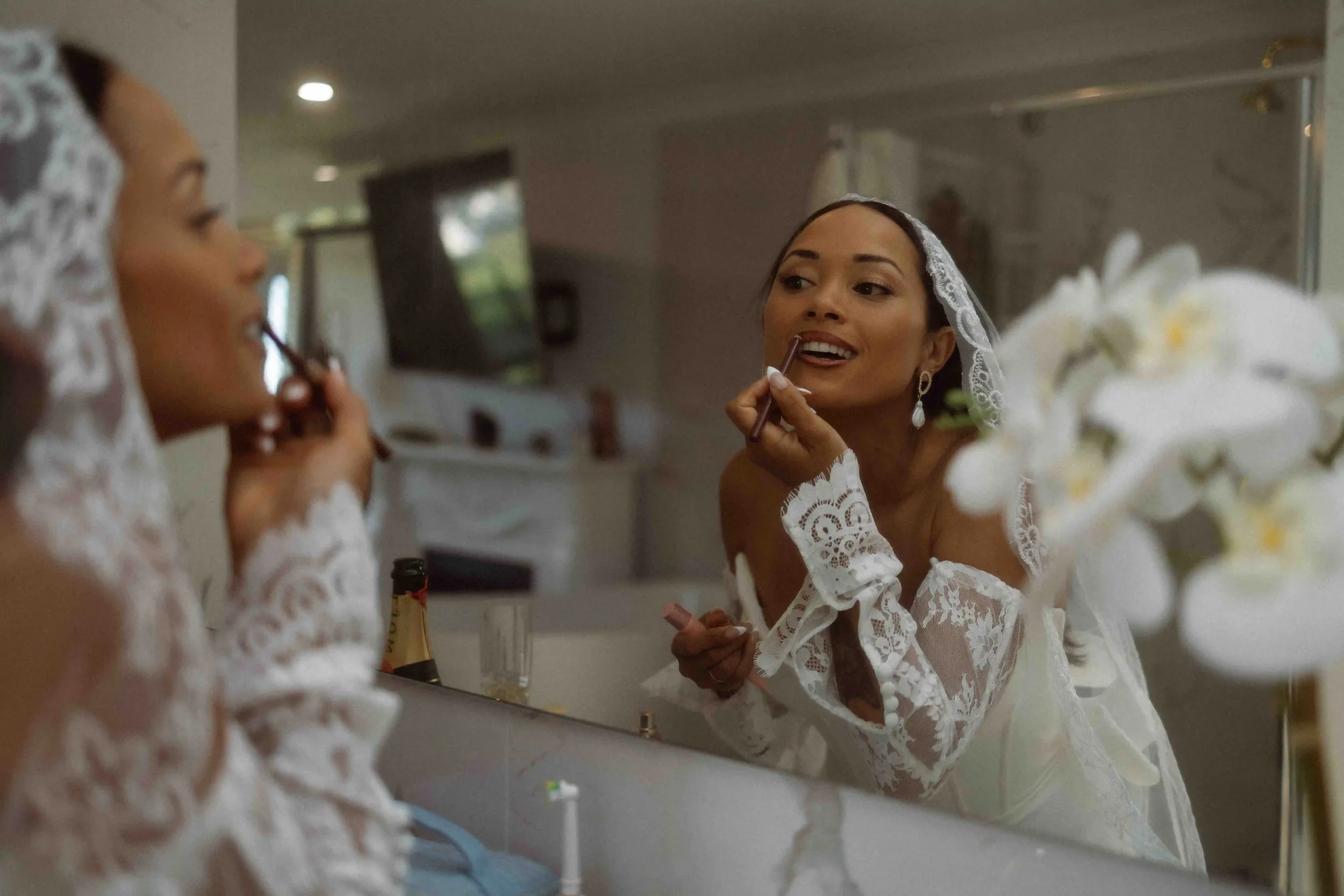 A woman in a white lace dress and veil is applying makeup in front of a mirror, while another woman in similar attire looks on nearby; a bouquet of white flowers is in the foreground.