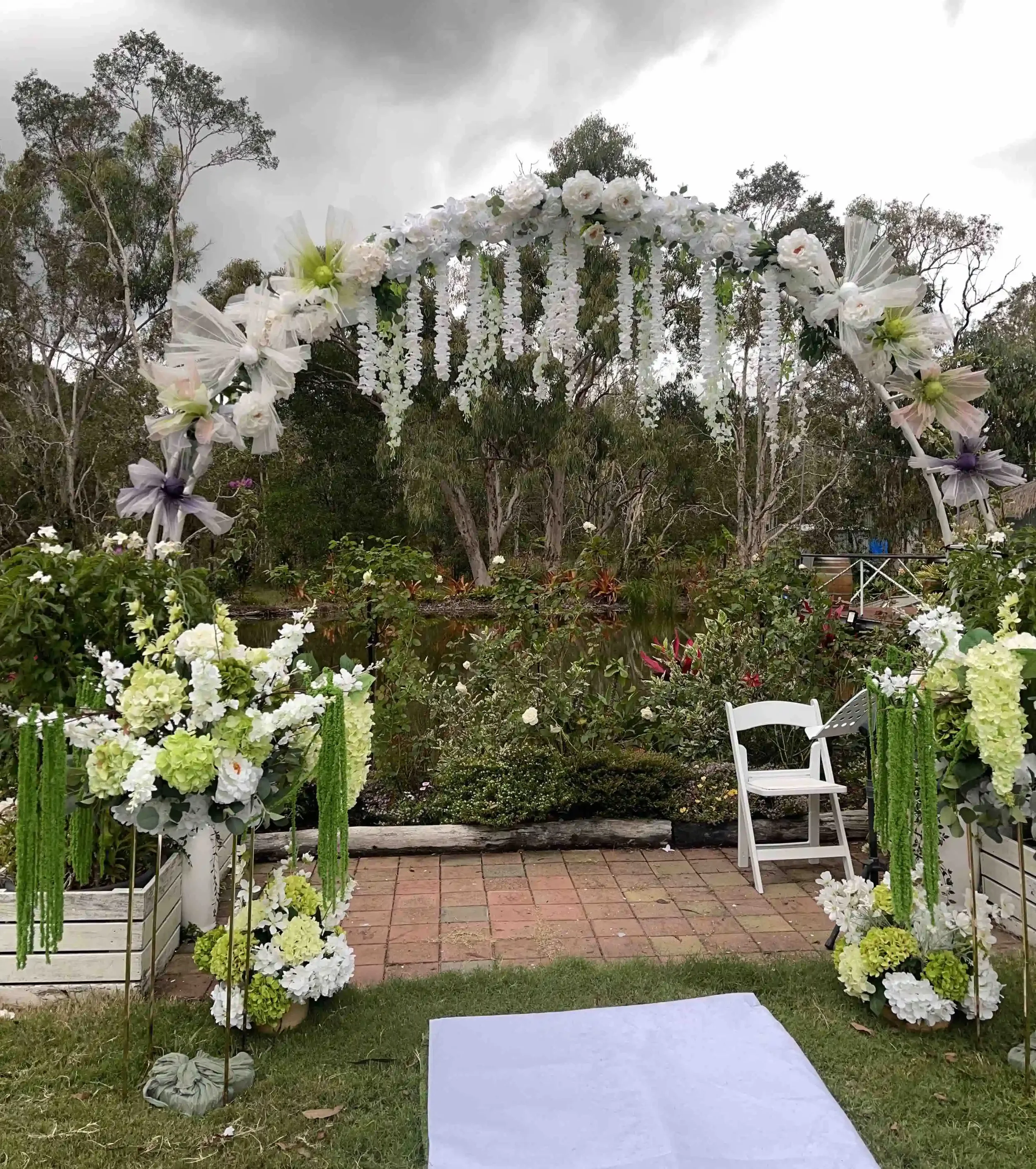 Outdoor wedding arch decorated with white flowers and hanging floral garlands, set against a backdrop of trees and cloudy sky, with chairs nearby and a white aisle runner.