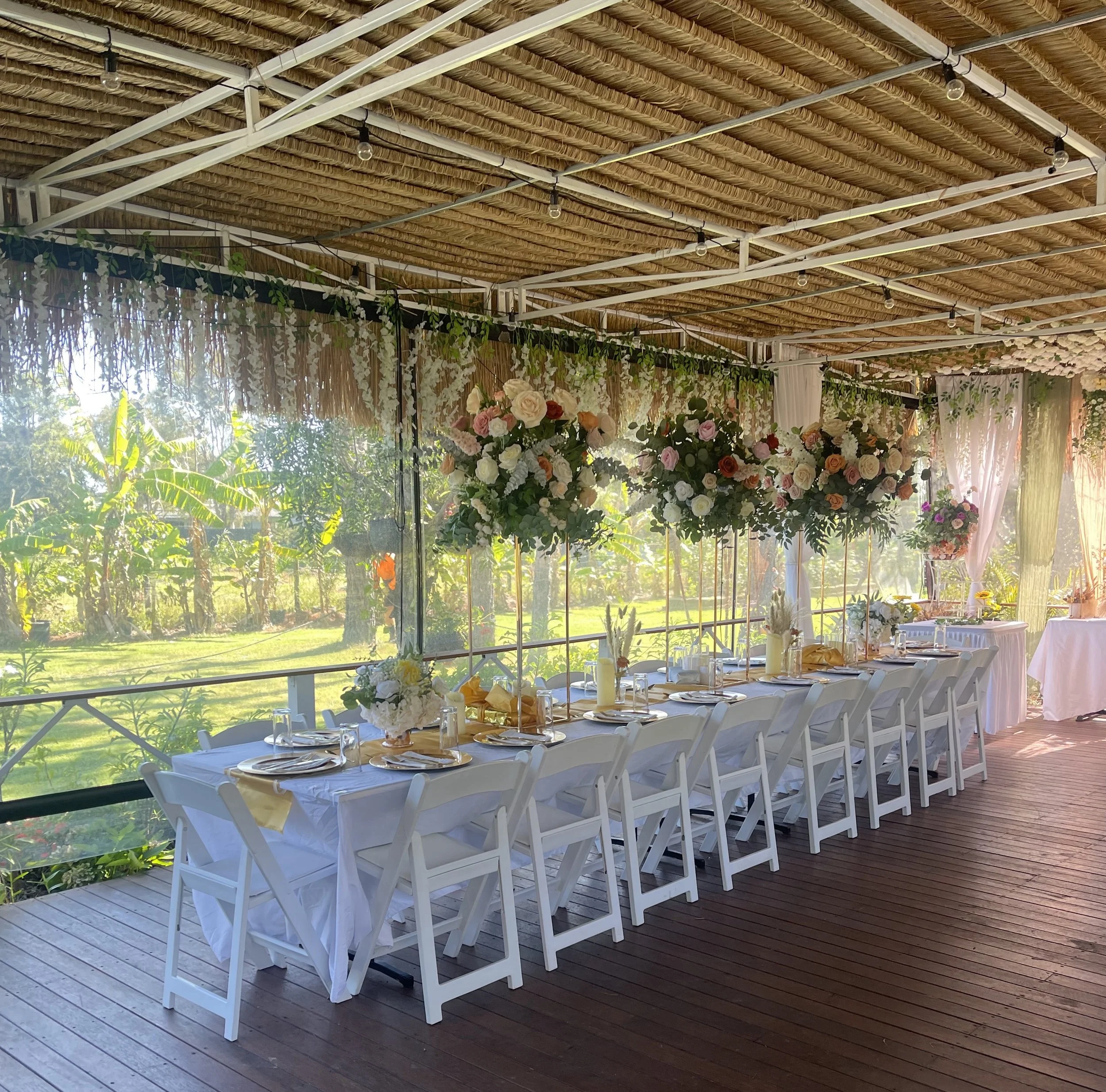 A beautifully decorated outdoor dining area with a long table, white chairs, and elegant floral centerpieces, set against a lush green garden backdrop with sunlight streaming through the windows.