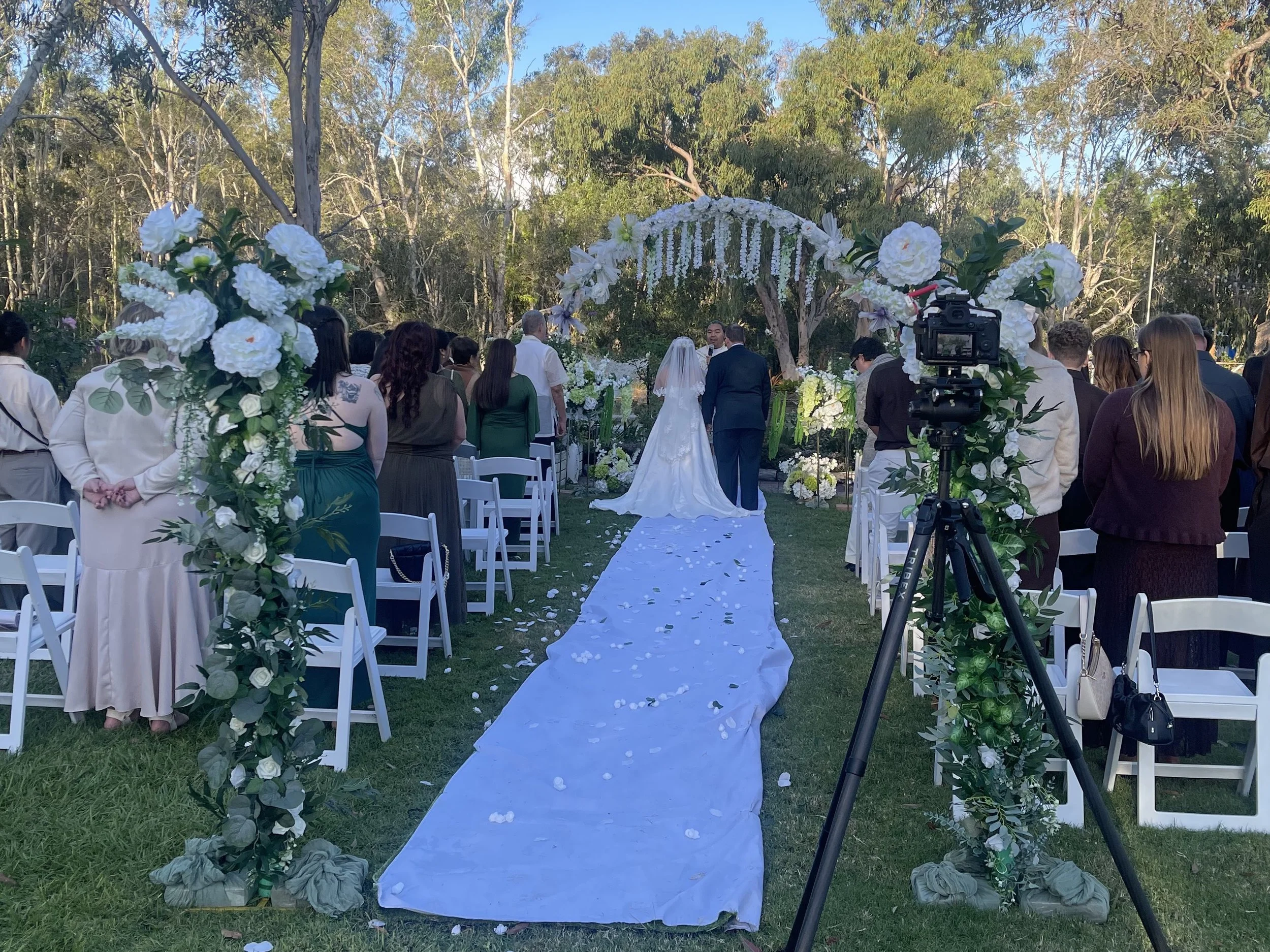 Outdoor wedding ceremony with an aisle decorated with flower arrangements, and a bride and groom standing at the altar under a floral arch. Guests seated on either side of the aisle overlook the ceremony, with trees and clear sky in the background.
