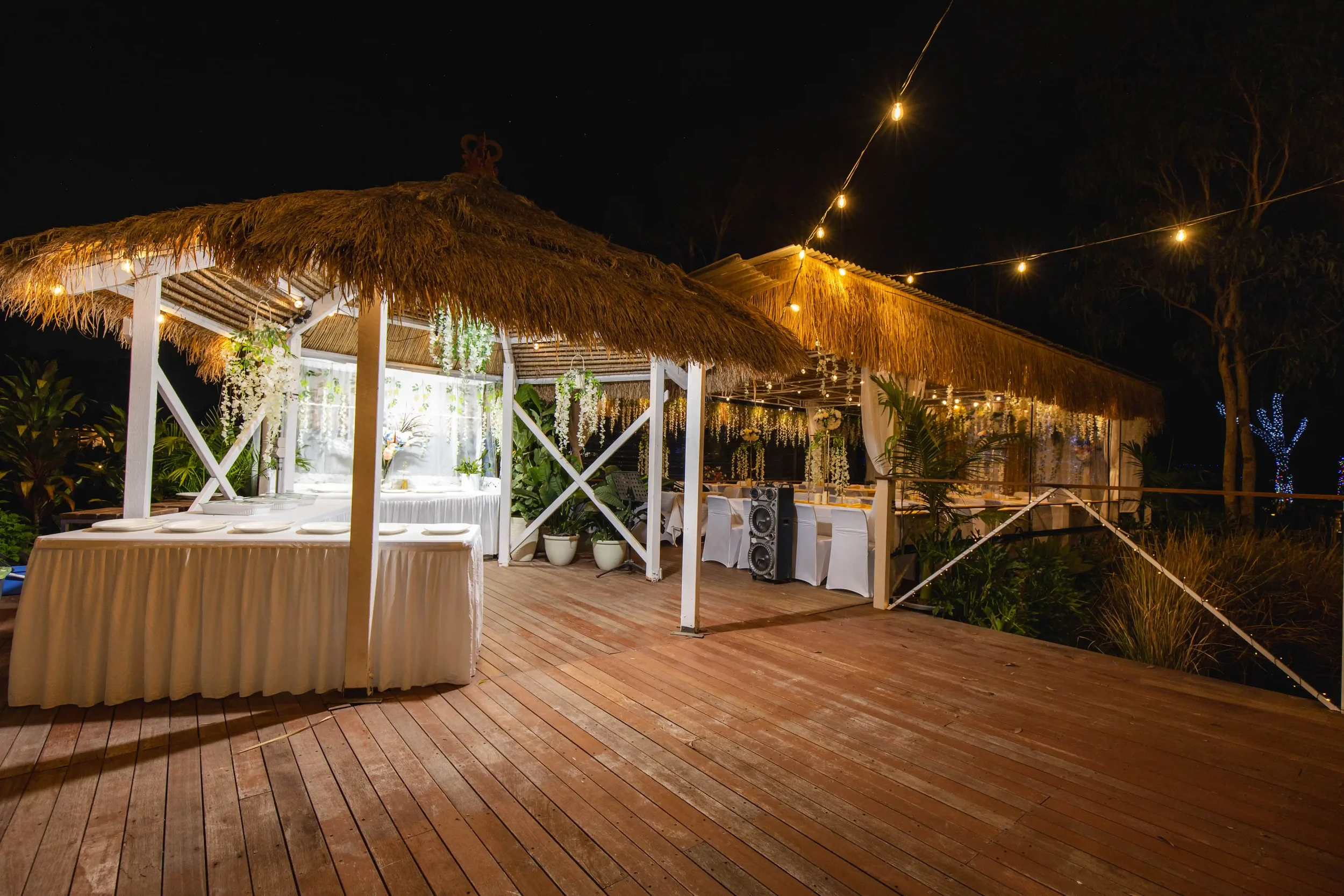 Nighttime outdoor event setup with thatched roof tents, string lights, white chairs, floral decorations, potted plants, and a sound system on a wooden deck.