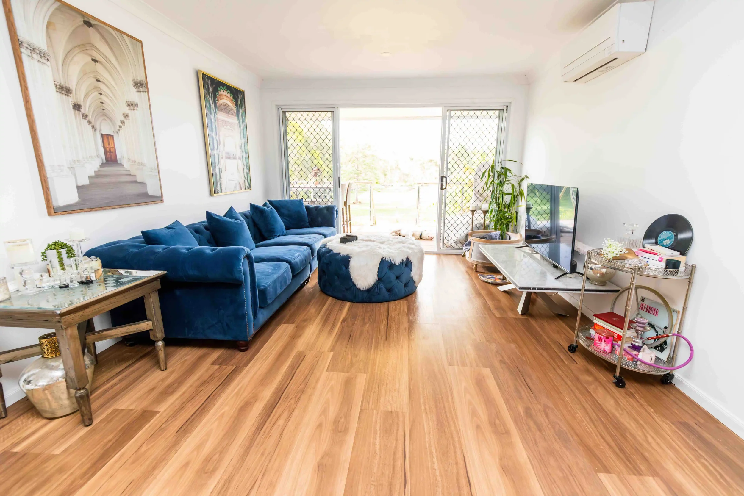 Living room with wooden floors, a large blue sofa with multiple cushions, a round ottoman with a white fur throw, and a glass sliding door leading to a balcony. There are paintings on the wall, a TV on a stand, a small table with plants, and children's toys.