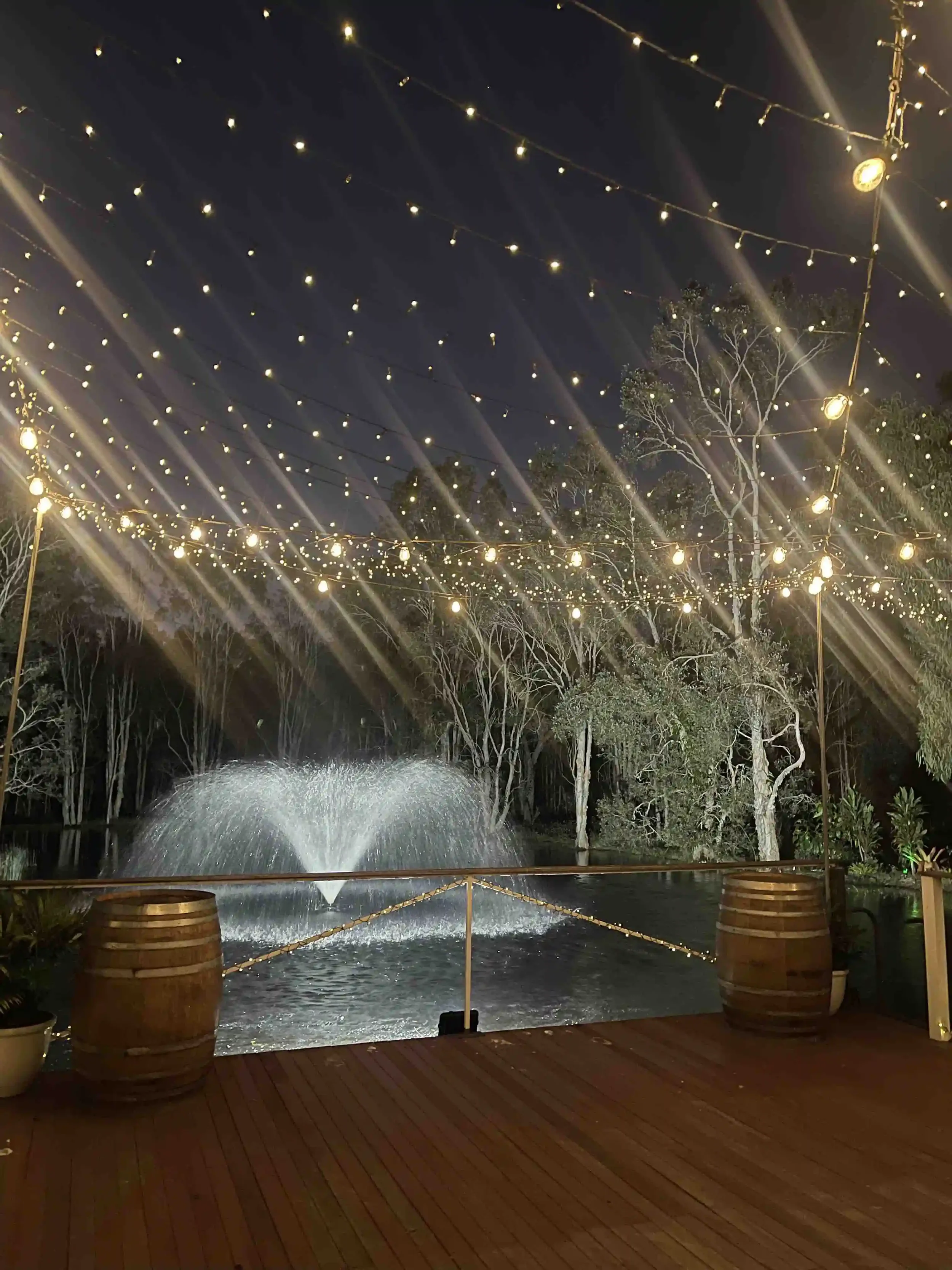 Night scene with string lights hanging above a pond with a fountain. Trees surround the pond, and there are wooden barrels on the patio.