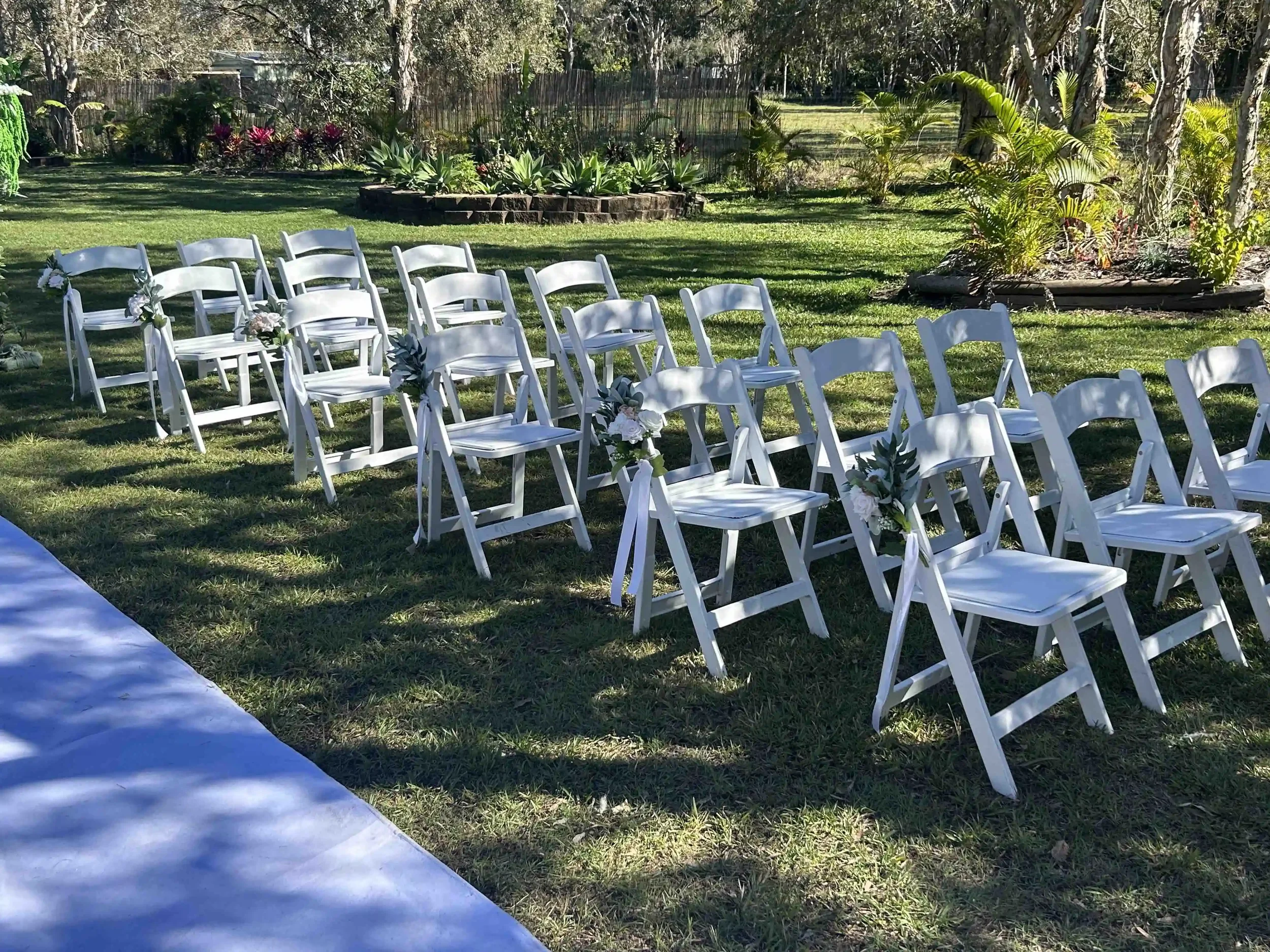 Outdoor wedding setup with white chairs decorated with flowers, arranged on a grassy lawn surrounded by trees and plants.