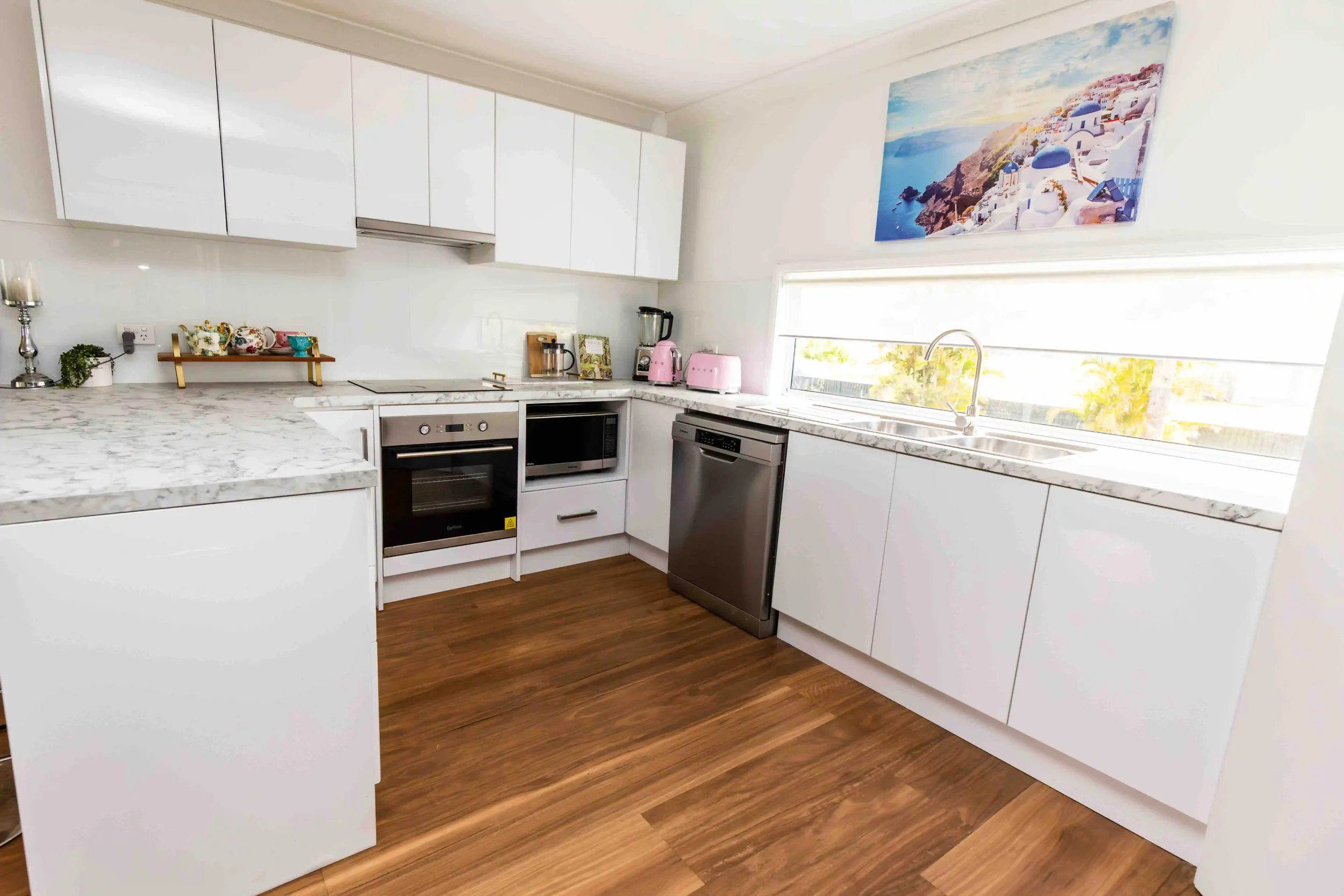 Modern kitchen with white cabinets, marble countertops, stainless steel oven, dishwasher, microwave, pink appliances, decorative cups and a large landscape painting of a Greek island scene on the wall.