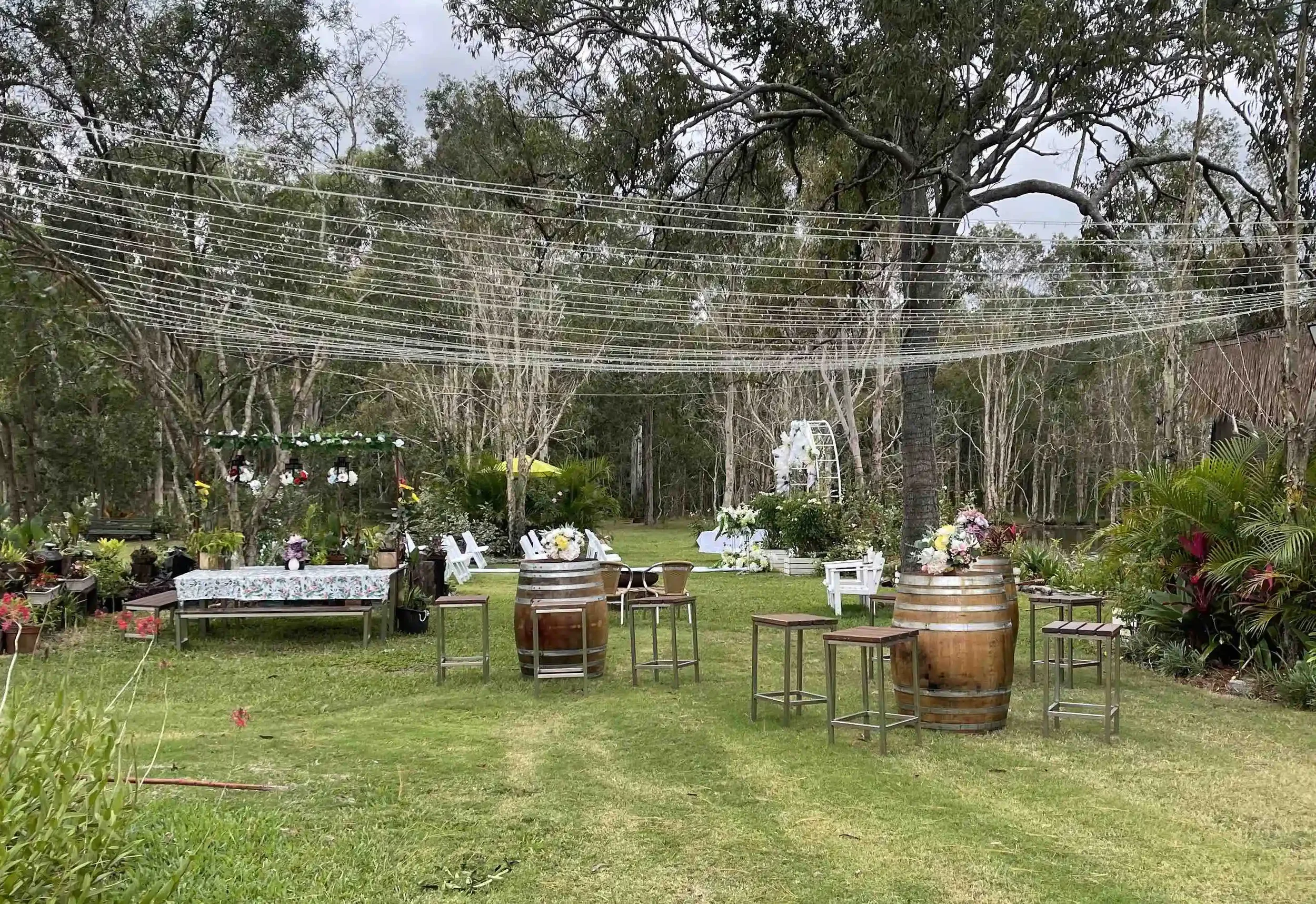 Outdoor garden setup with tables, chairs, floral arrangements, and string lights, suggesting a celebration or event space in a lush, wooded area.