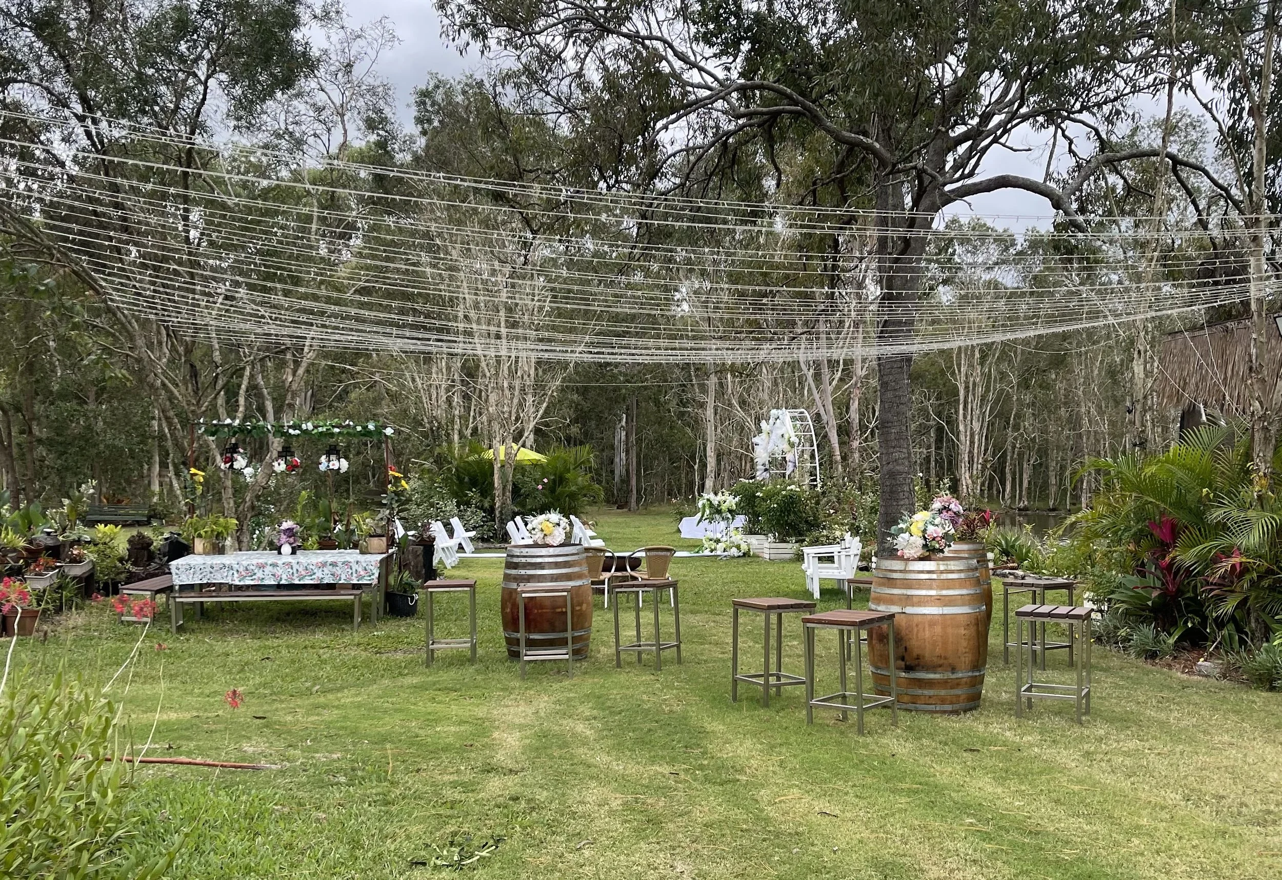 Outdoor garden setup with wine barrels, chairs, floral decorations, and string lights, surrounded by trees and greenery.