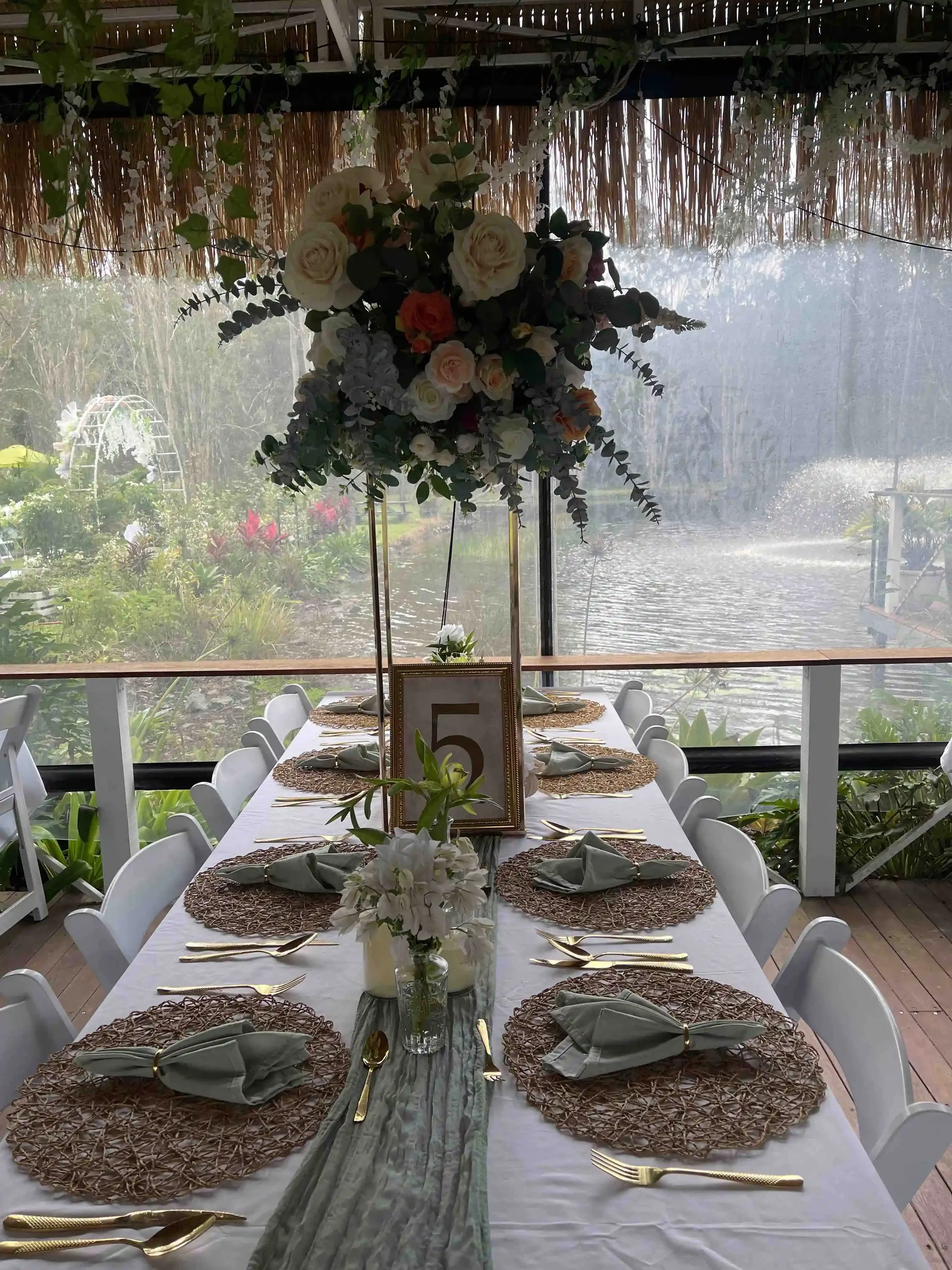 A long banquet table decorated for a celebration, with woven placemats, gold utensils, folded gray napkins, and a floral centerpiece with white and orange roses. The table is set in front of a large window overlooking a garden and pond with a fountai