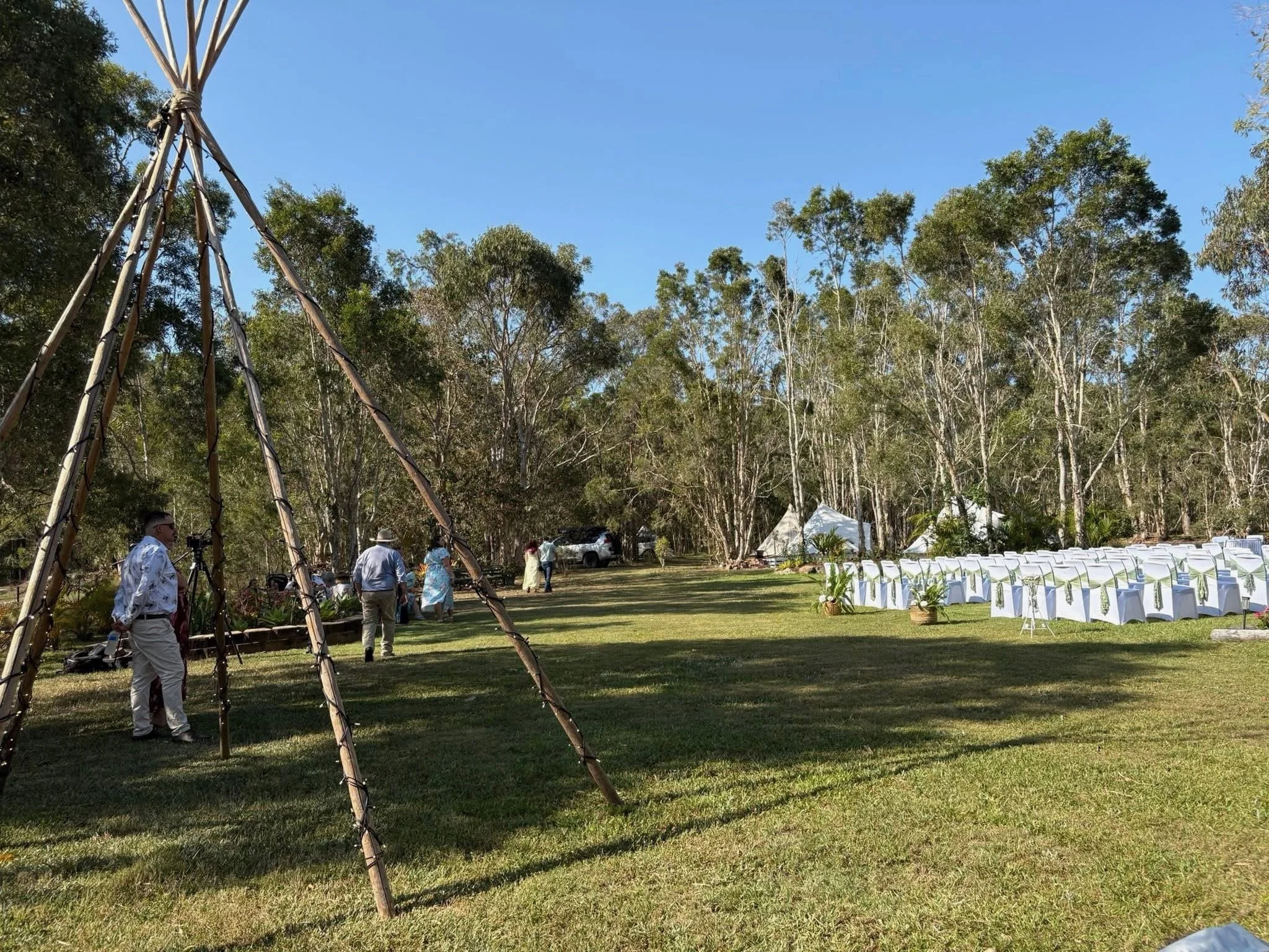 Outdoor wedding setup with chairs decorated with white covers and green sashes, white tents among trees, and a naked teepee in the foreground on a sunny day.