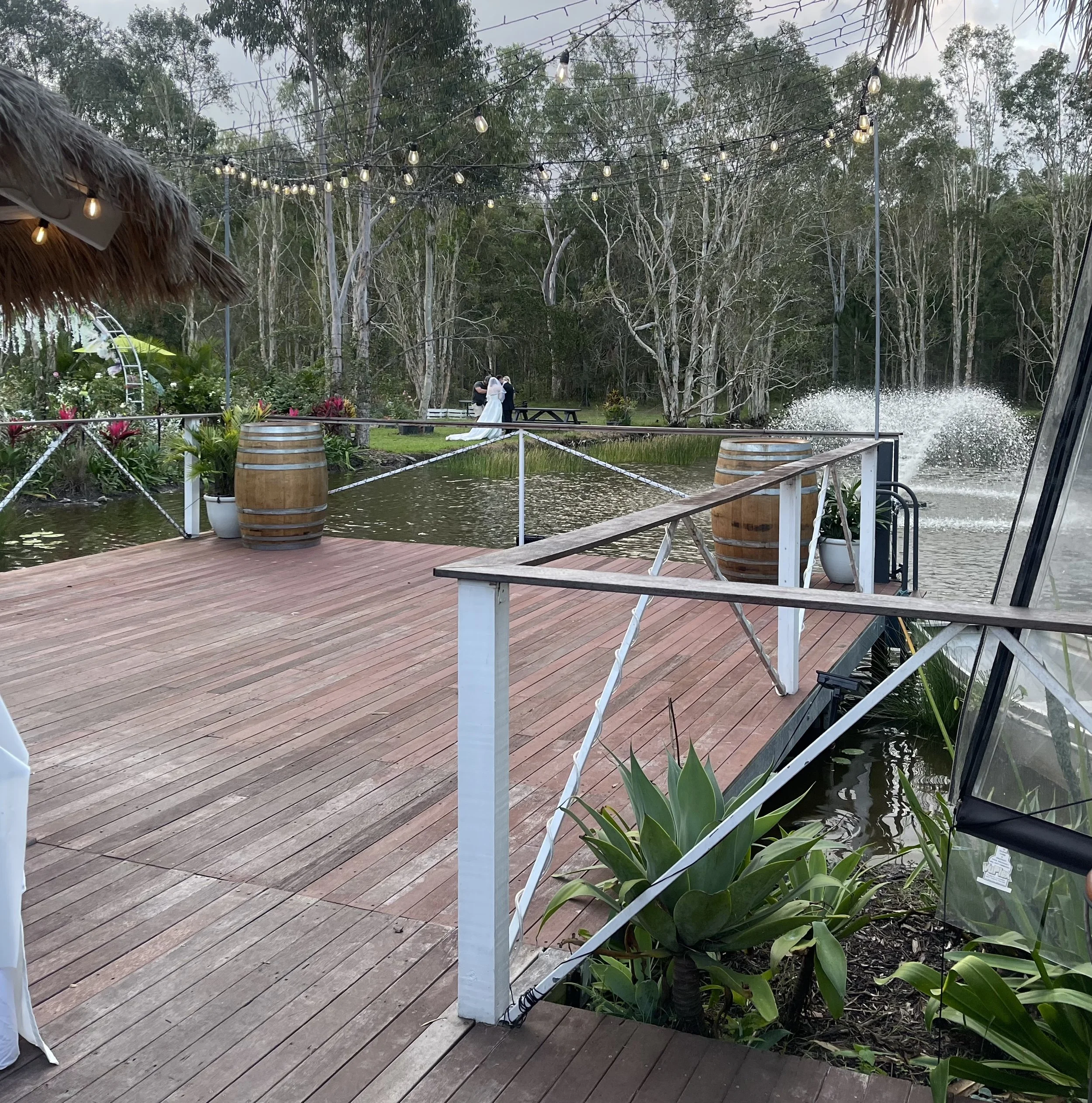 Outdoor scene with string lights hanging above a wooden deck surrounded by trees and greenery. A couple dressed in wedding attire, with a bride in a white gown and a groom in a dark suit, stand by a bench, at a wedding.