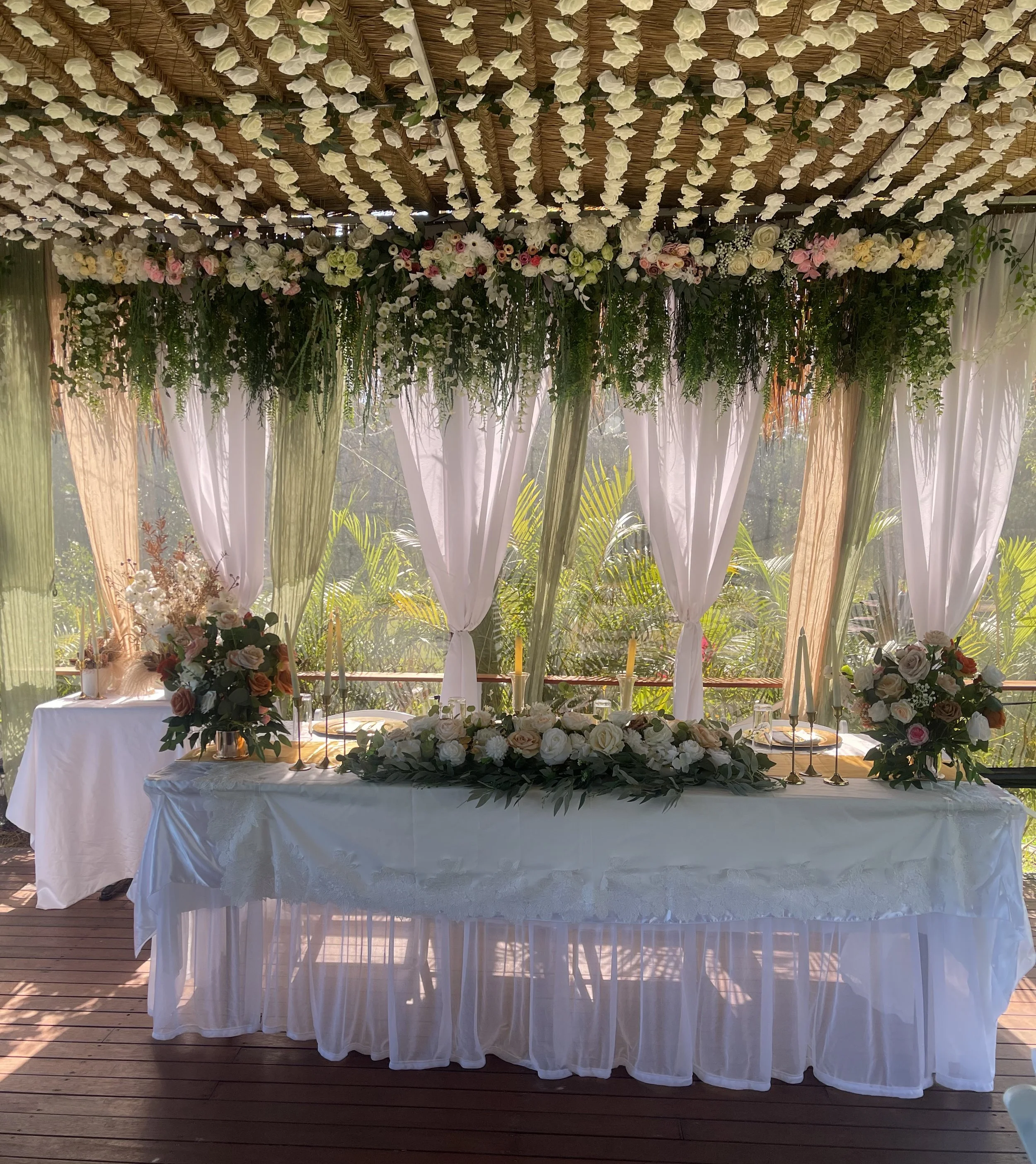 Decorated outdoor table with floral arrangements, candles, and draped curtains under a floral canopy, suitable for a wedding or celebration.