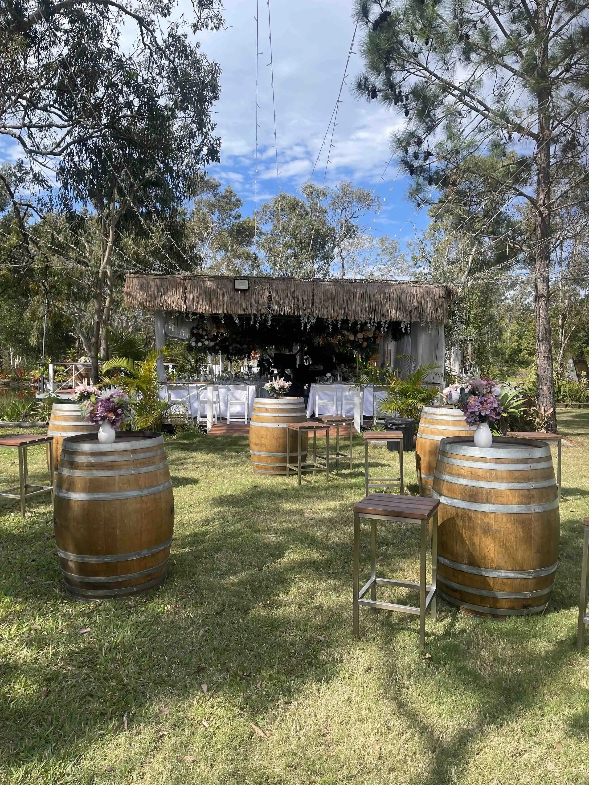 Outdoor wedding setup with a small stage, decorated with flowers, under a thatched roof, surrounded by trees, with tables made from wine barrels with flower arrangements, and chairs arranged for guests.