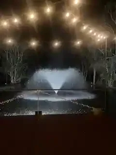 Nighttime scene with string lights overhead and a fountain in the shape of a heart with water spraying upward
