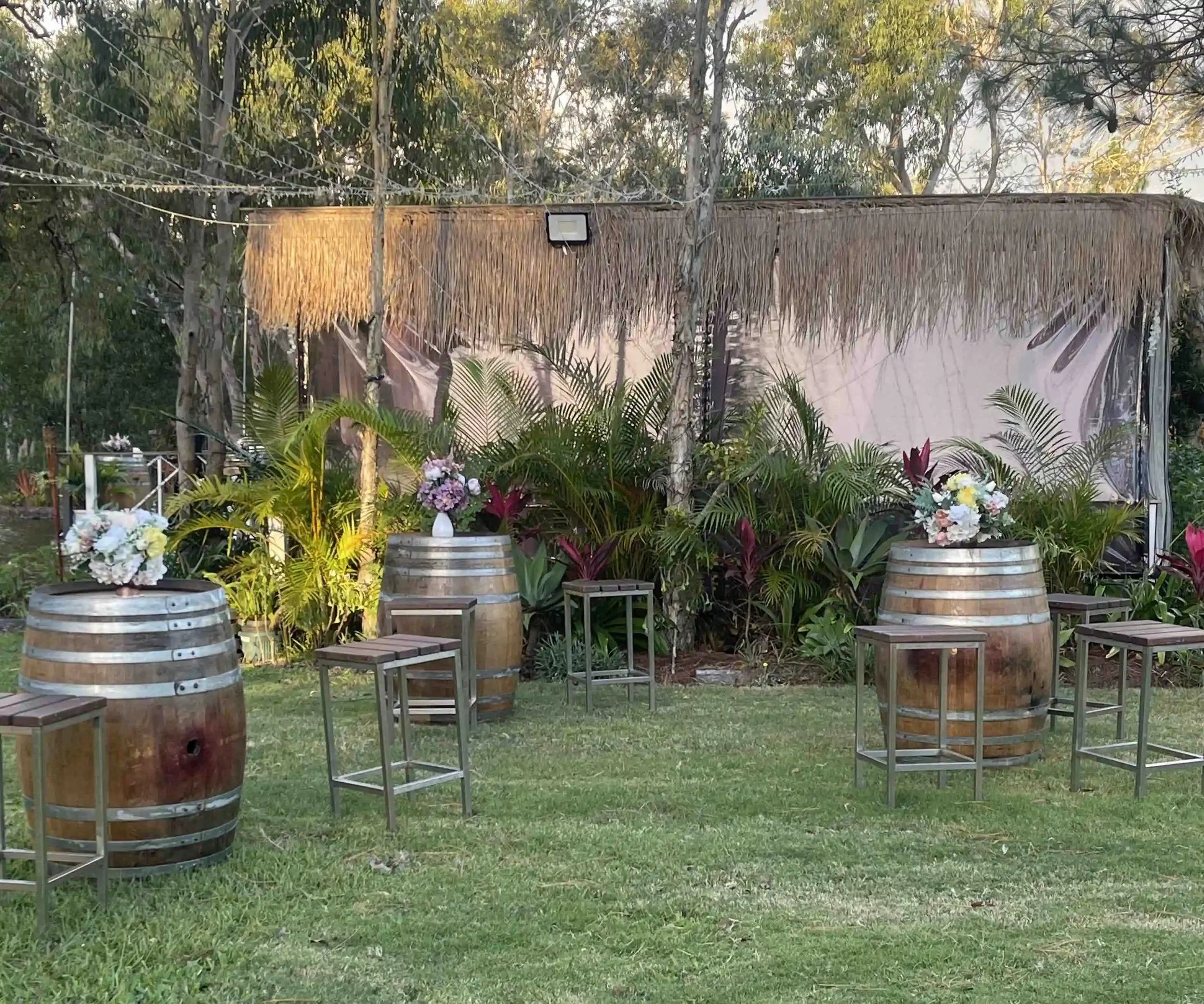 Outdoor decor scene featuring wooden barrels, small wooden stools, lush green plants, and floral arrangements, with a thatched roof structure and string lights in the background.