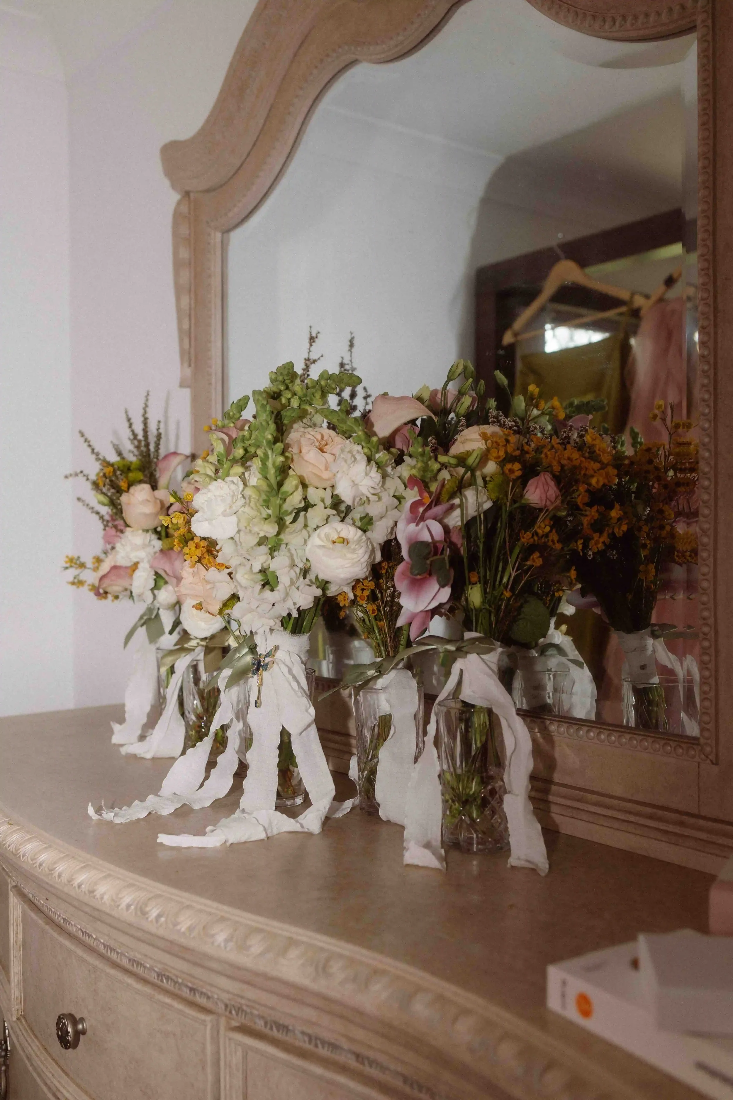 A wooden dresser with a large mirror reflecting a bouquet of pink, white, and yellow flowers in glass vases, and a cardboard box on the right side.