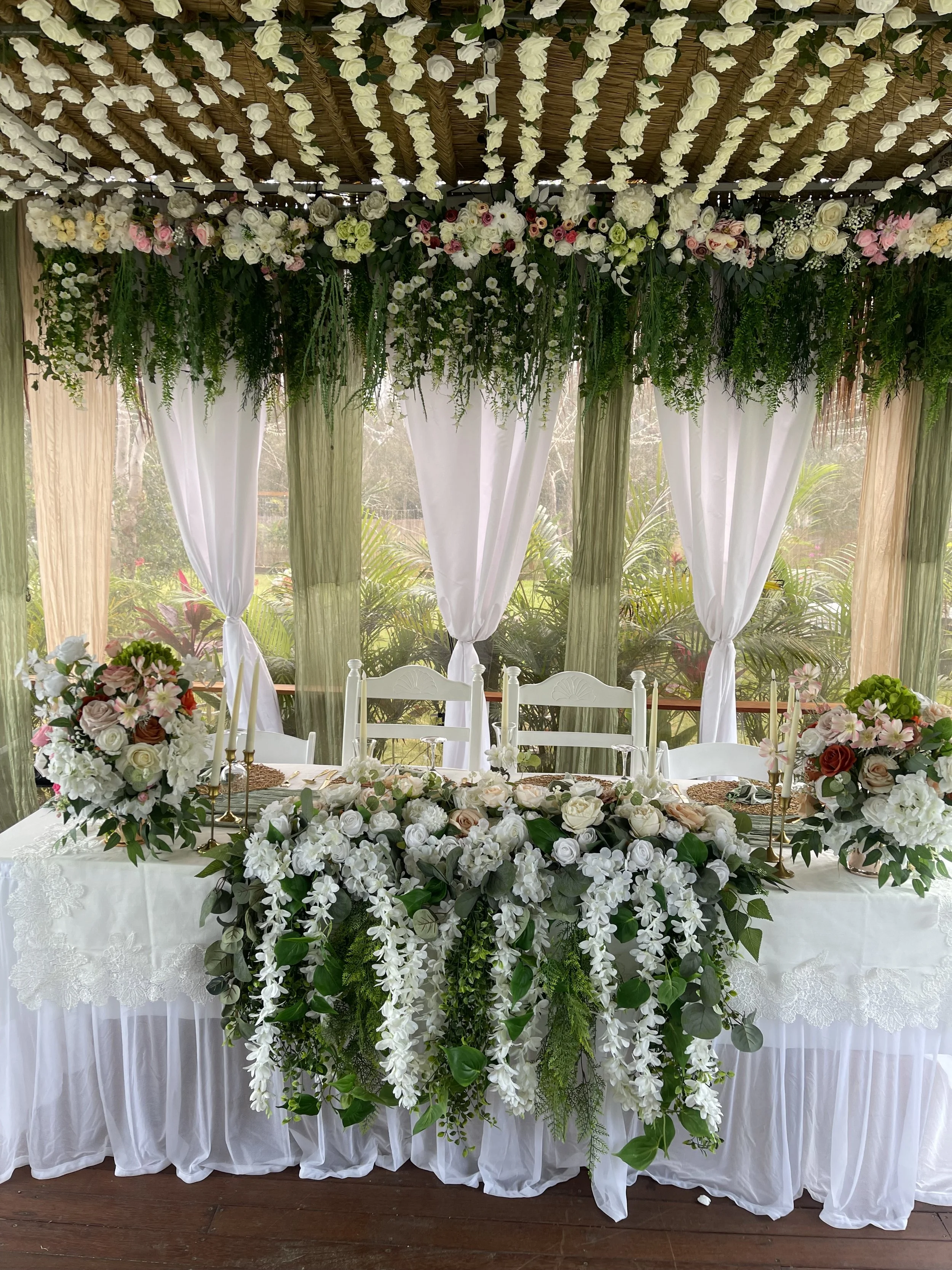 Wedding or event decoration with a long white draped table, floral arrangements in white, blush pink, and mauve, and a lush greenery centerpiece. The backdrop features hanging white flowers, and sheer white curtains are tied back on either side.