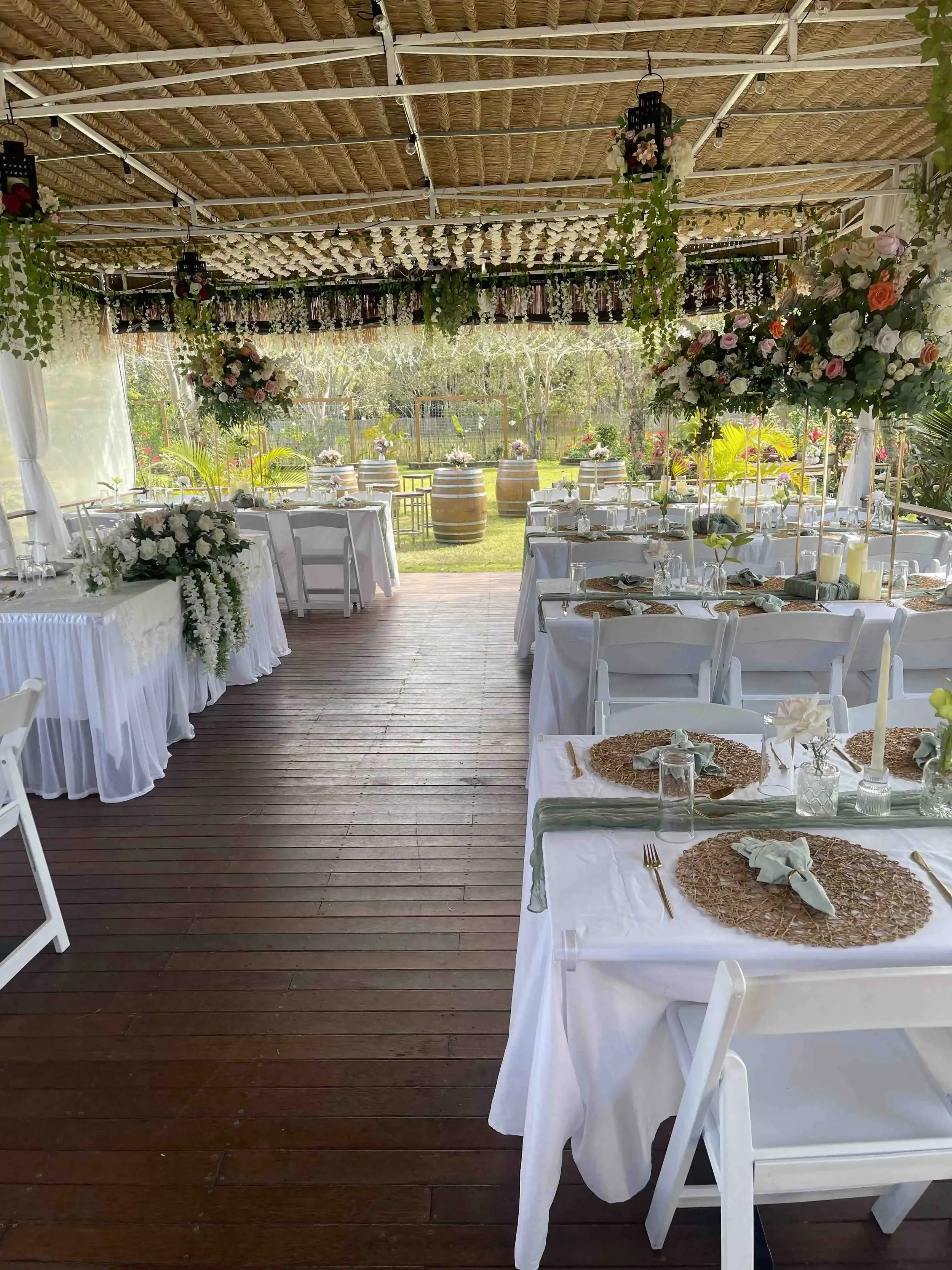 A decorated outdoor wedding reception space with tables dressed in white tablecloths and set with plates, glasses, and napkins. There are floral centerpieces and candles on the tables, and floral arrangements hang from the ceiling. The space has a wooden floor and is open to a lush green outdoor area with trees and foliage visible in the background, and electric string lights hanging from the ceiling.