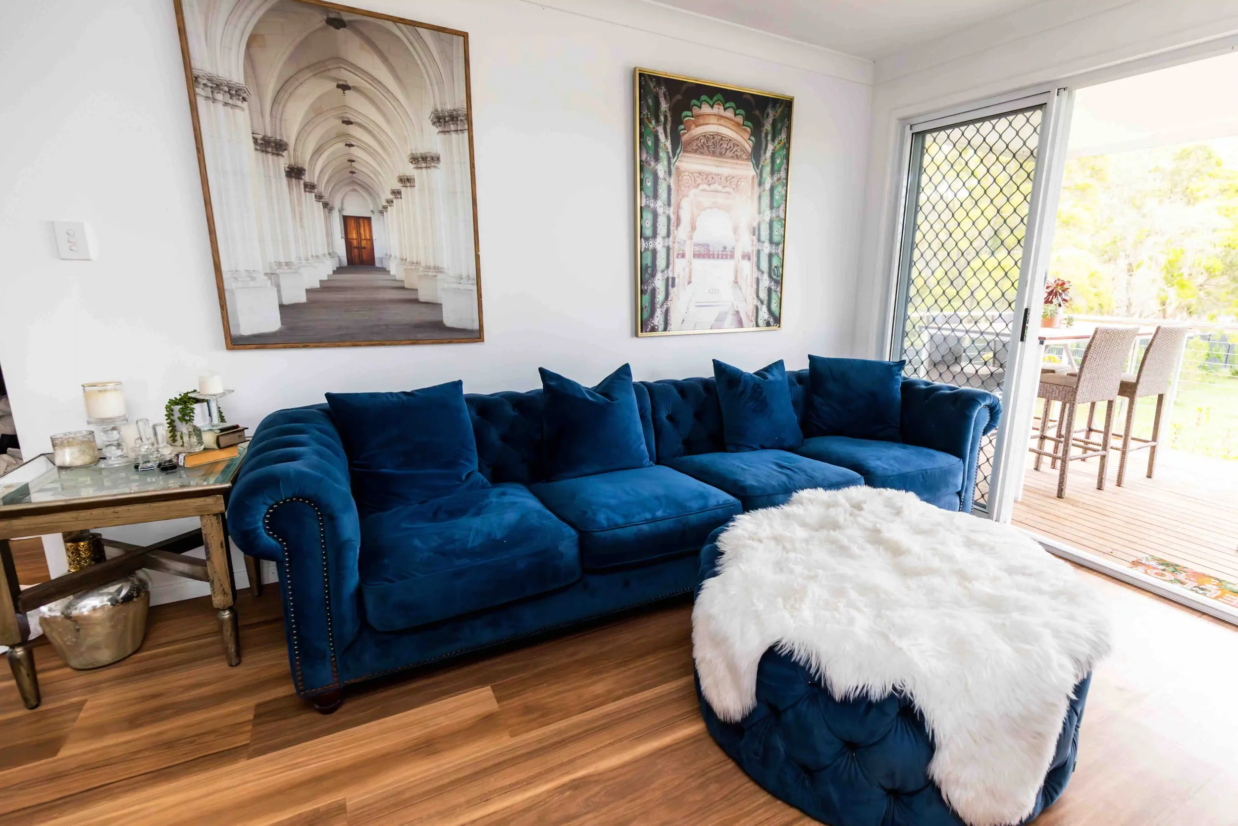 Living room with a large blue velvet tufted sofa, two art pieces on the wall, a sliding glass door leading to a balcony, and a white fluffy ottoman.