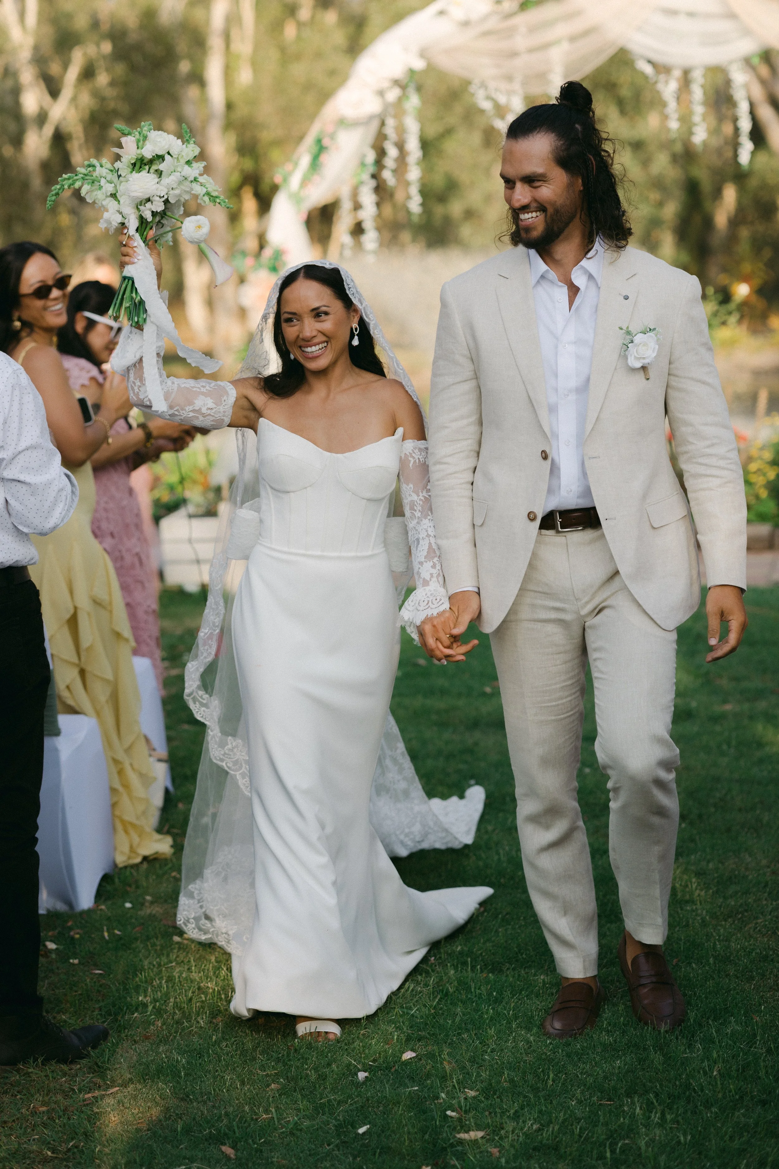 A newly married couple walking hand-in-hand outdoors at a wedding reception. The bride is holding a bouquet and smiling, wearing a white wedding dress and veil. The groom is dressed in a light-colored suit and is also smiling. Guests are clapping and