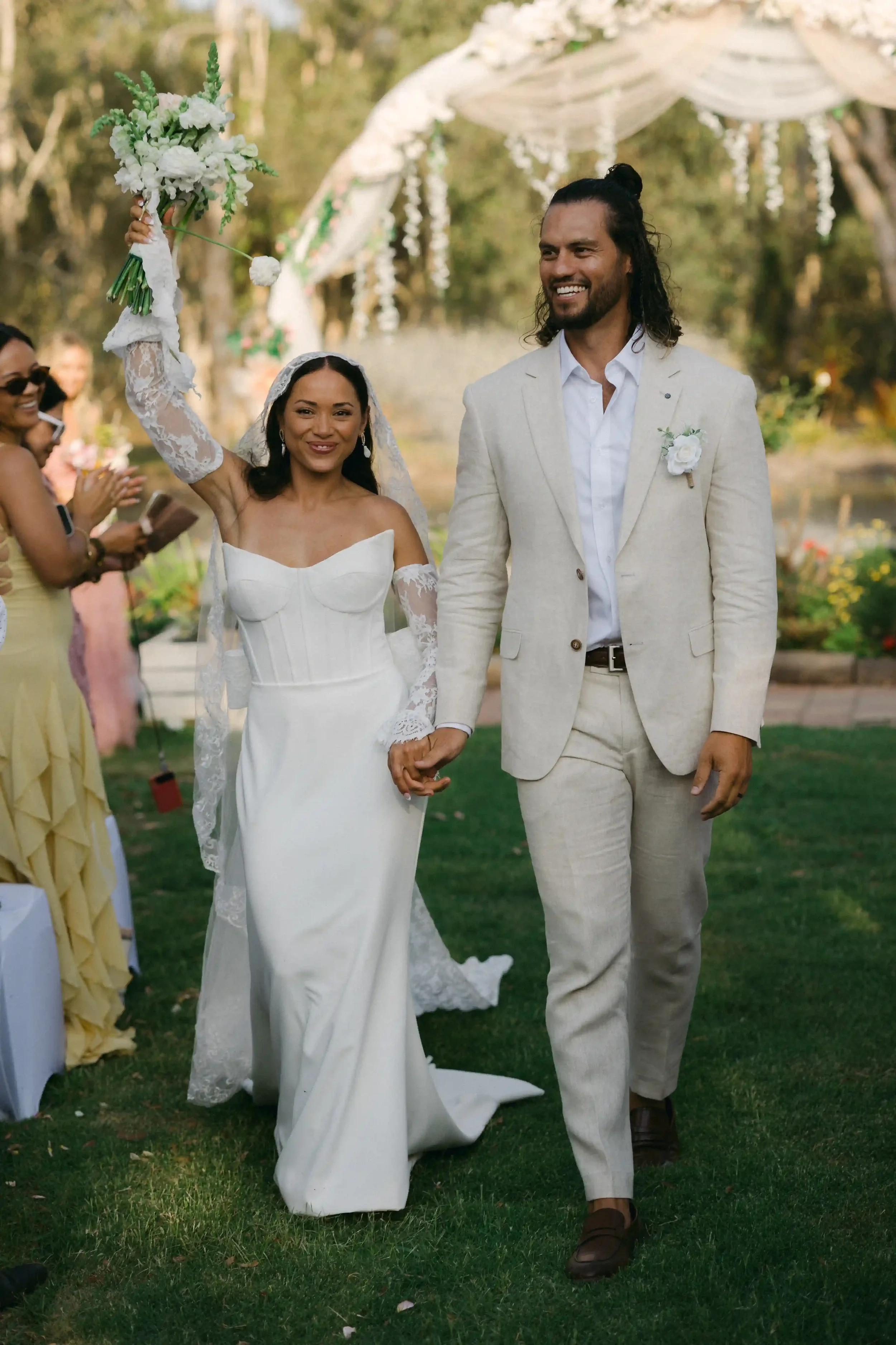 A couple holding hands and smiling at a wedding ceremony outdoors. The bride, wearing a white gown and veil, is raising a bouquet of white flowers. The groom is dressed in a light-colored suit. Guests are clapping in the background, and there is a de