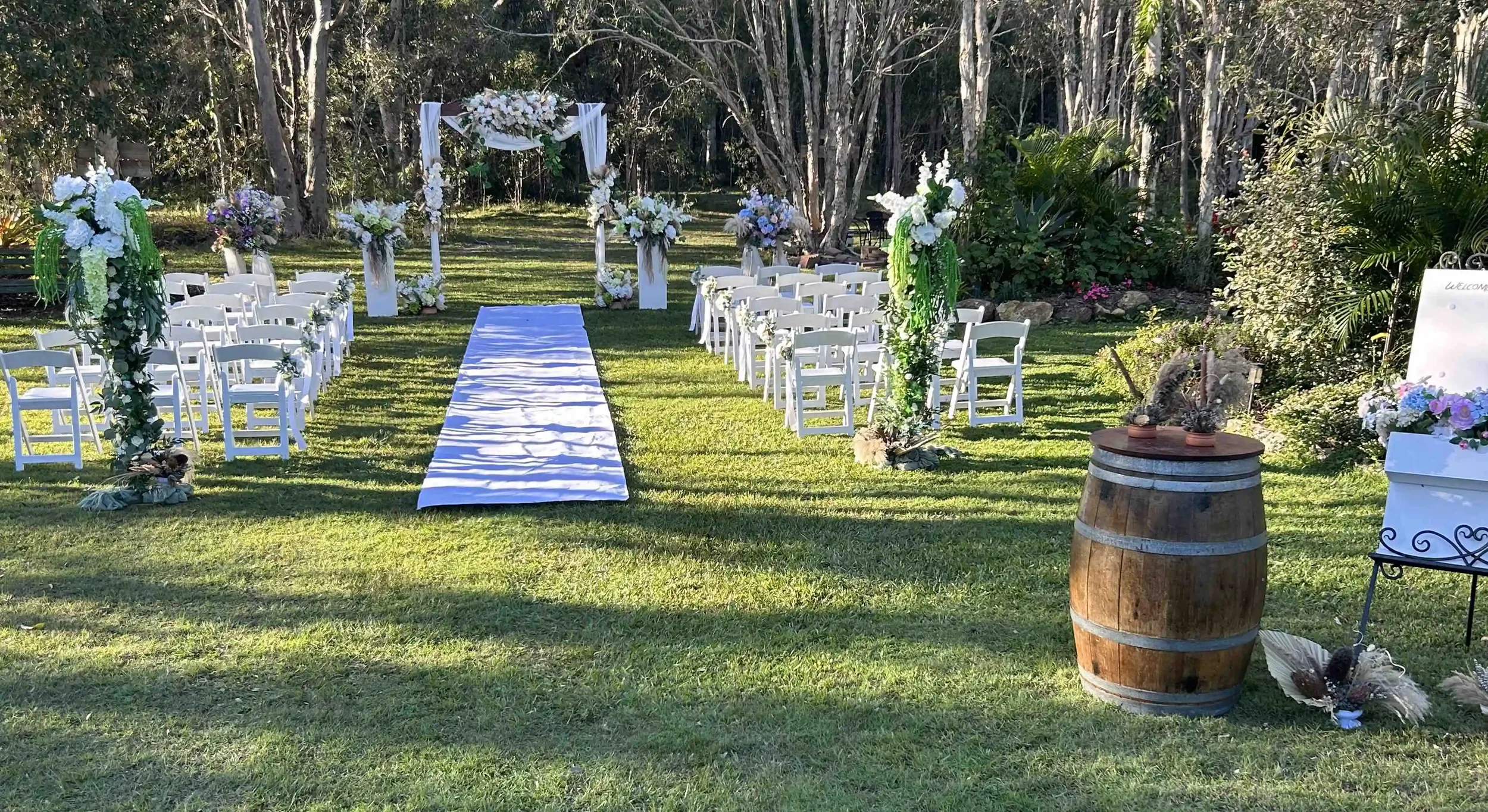 Outdoor wedding setup with white chairs lined on either side of a white aisle runner, floral arrangements along the aisle, a decorated arch at the front, and surrounded by trees and greenery.
