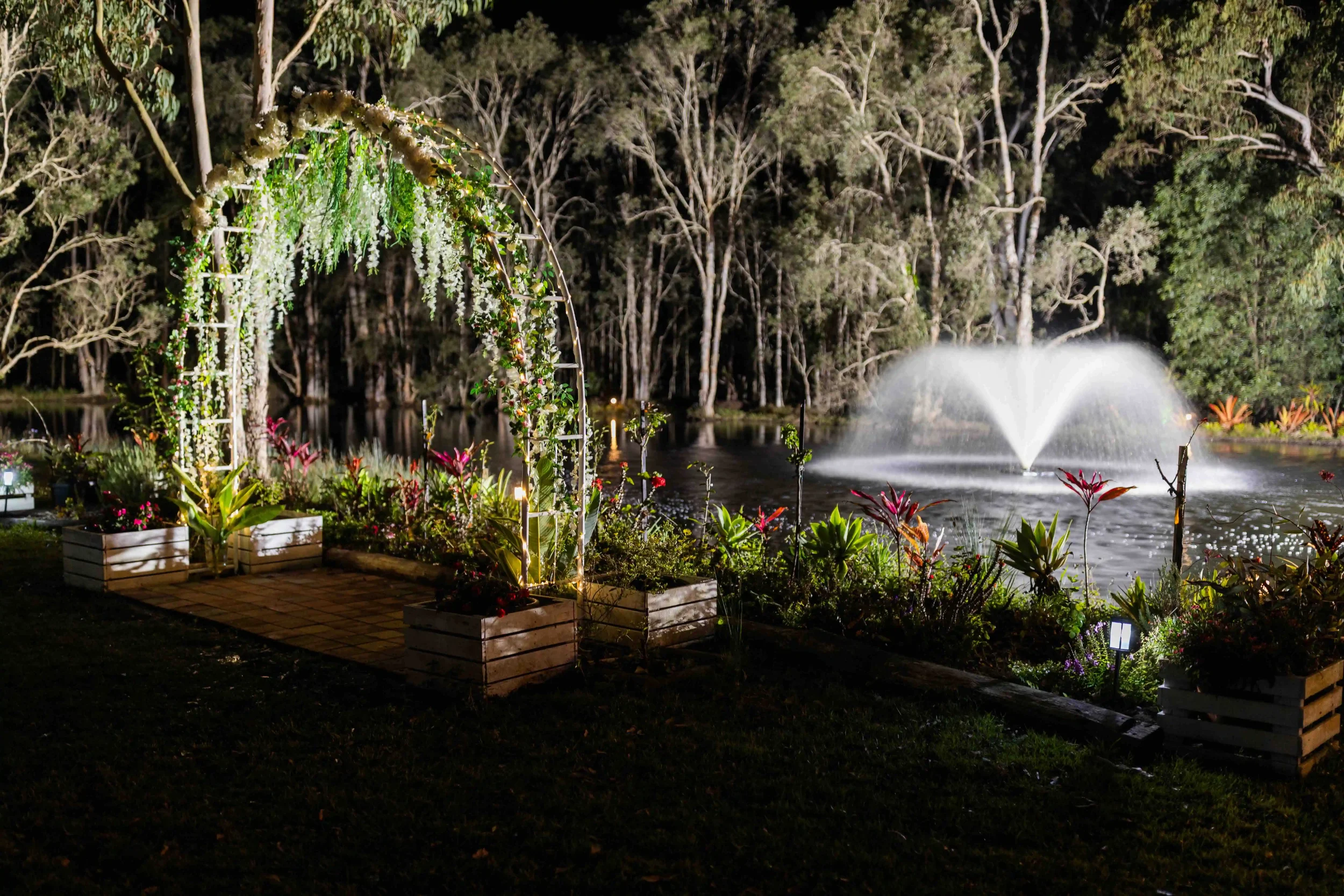 Nighttime outdoor scene with a decorative floral arch near a pond or lake, with a fountain spraying water in the background and trees surrounding the area.