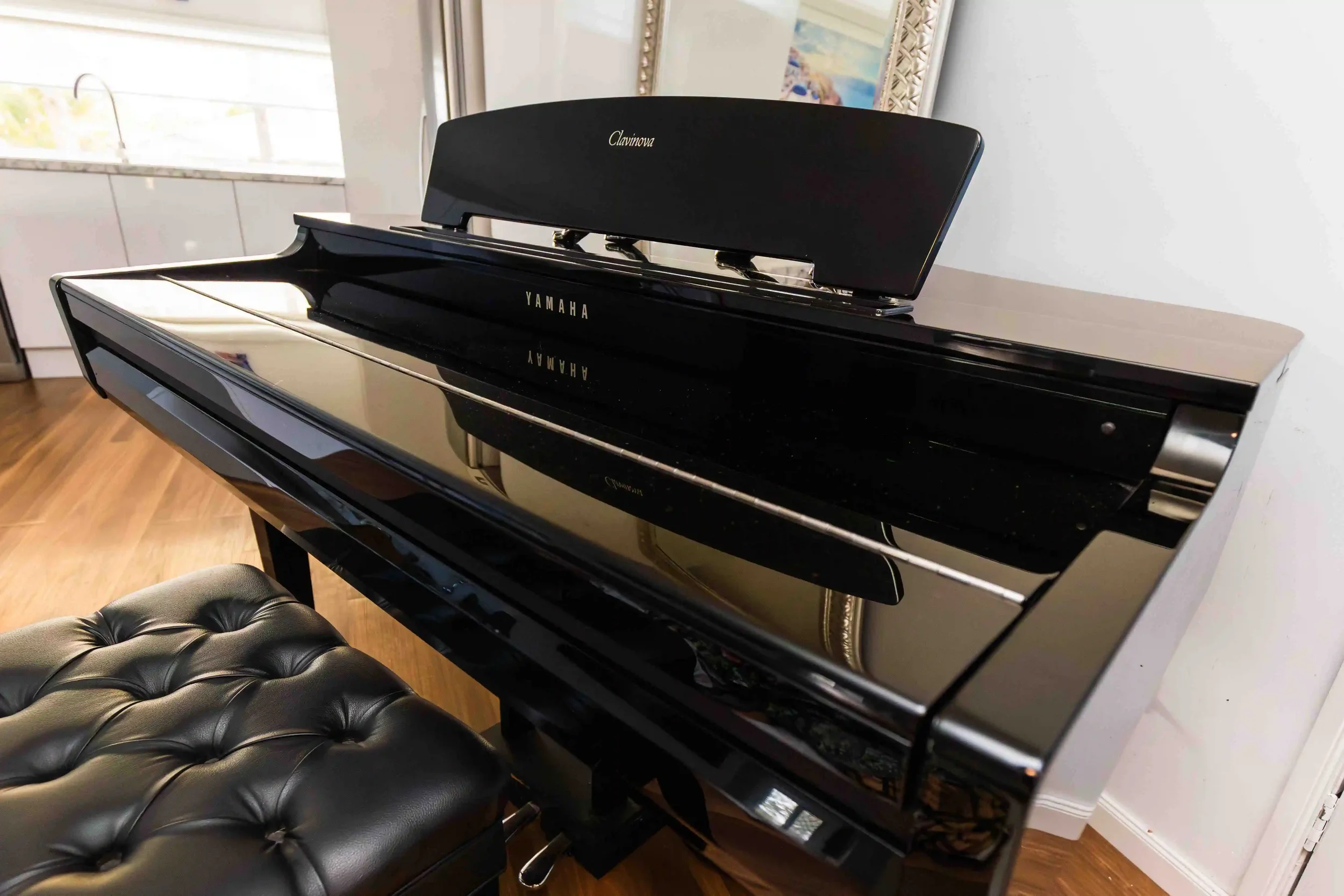 A black glossy Yamaha Clavinova digital piano with a matching black cushioned bench in a room with wooden floors and a framed painting hanging on a white wall.