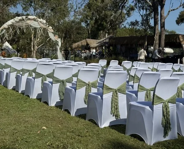 Rows of white wedding chairs with green sashes tied at the back, facing an arch decorated with flowers, outdoors in a garden setting.