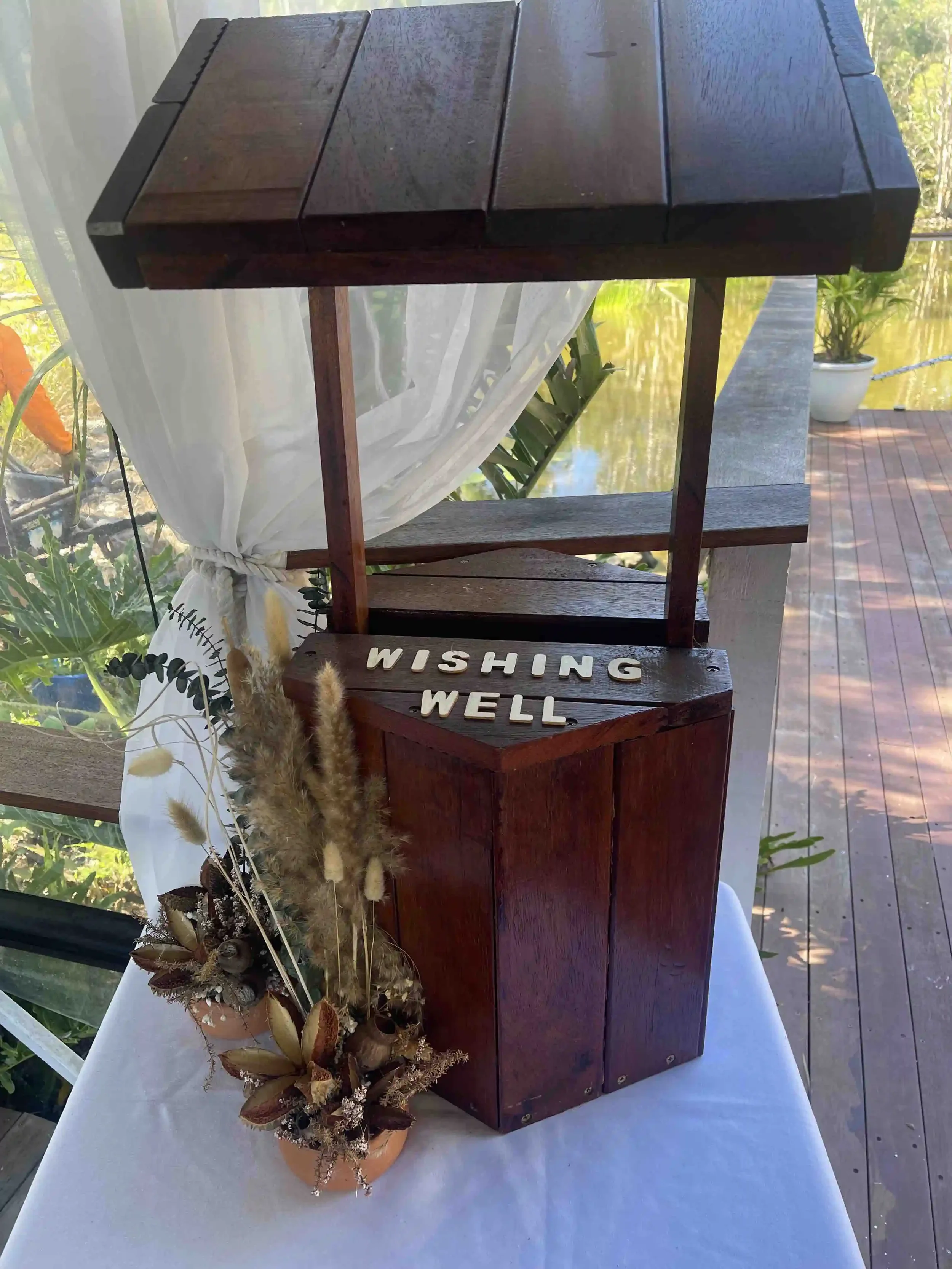 A wooden wishing well decoration with the words 'WISHING WELL' on it, set on a white table next to a potted plant with dried flowers, outdoors with a lake and trees in the background.