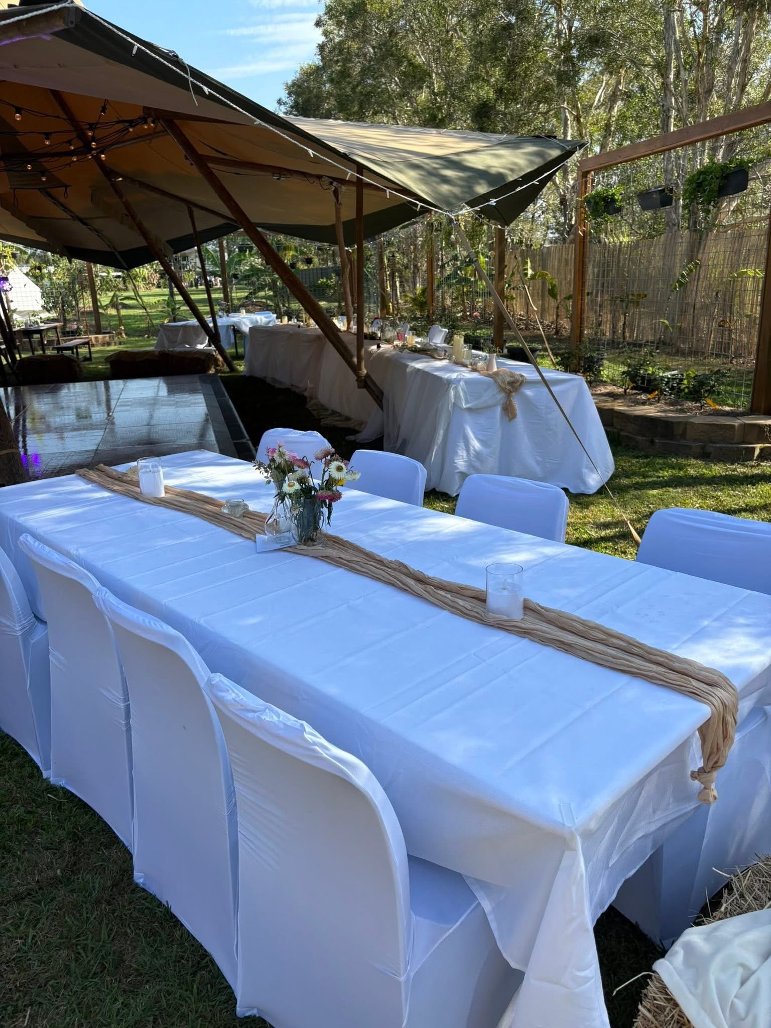 Outdoor event setup with a long white table covered with a white tablecloth and decorated with candles and a flower centerpiece, under a canopy in a backyard with trees.