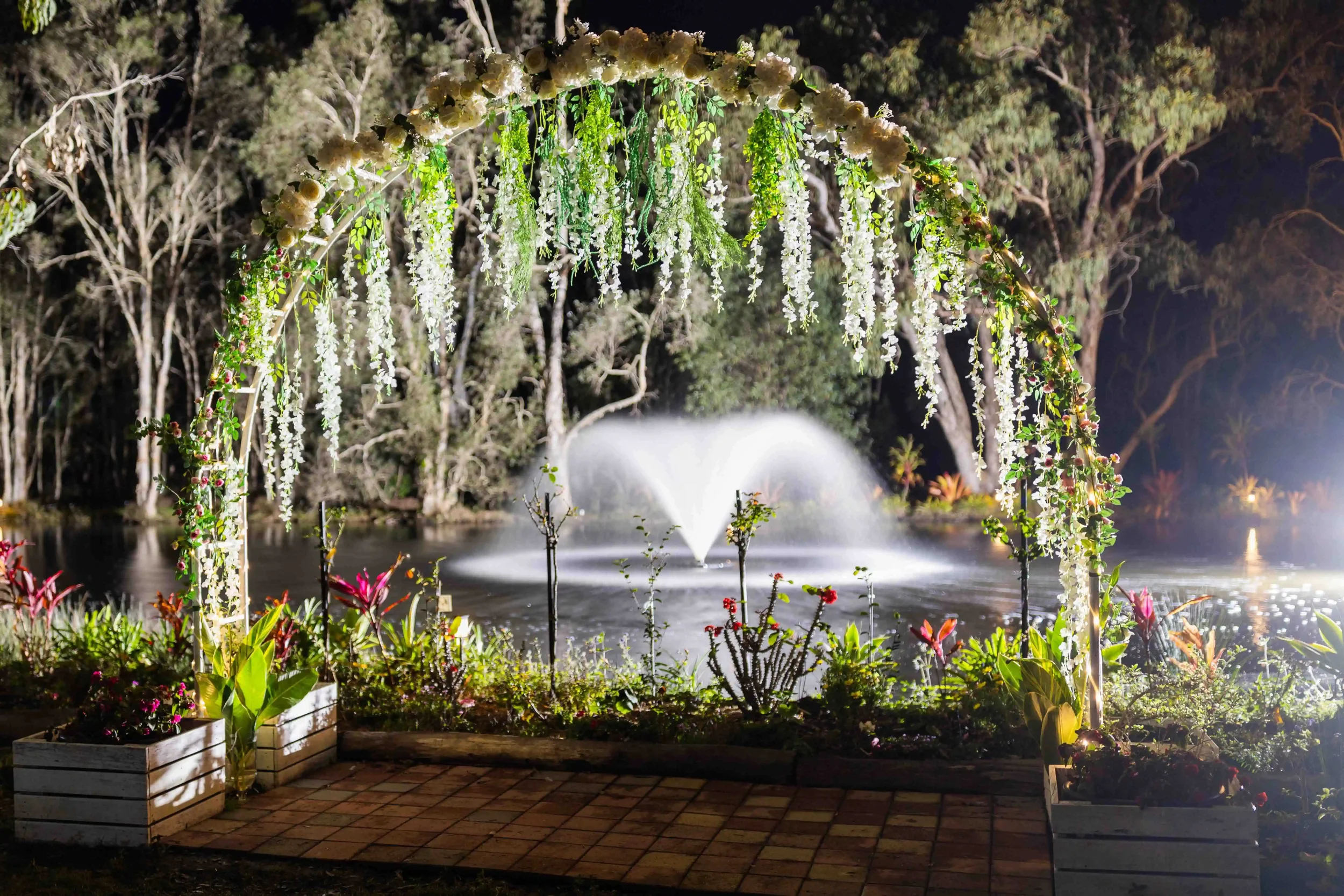 A floral wedding arch over a pond with fountain at night, surrounded by trees and colorful plants.