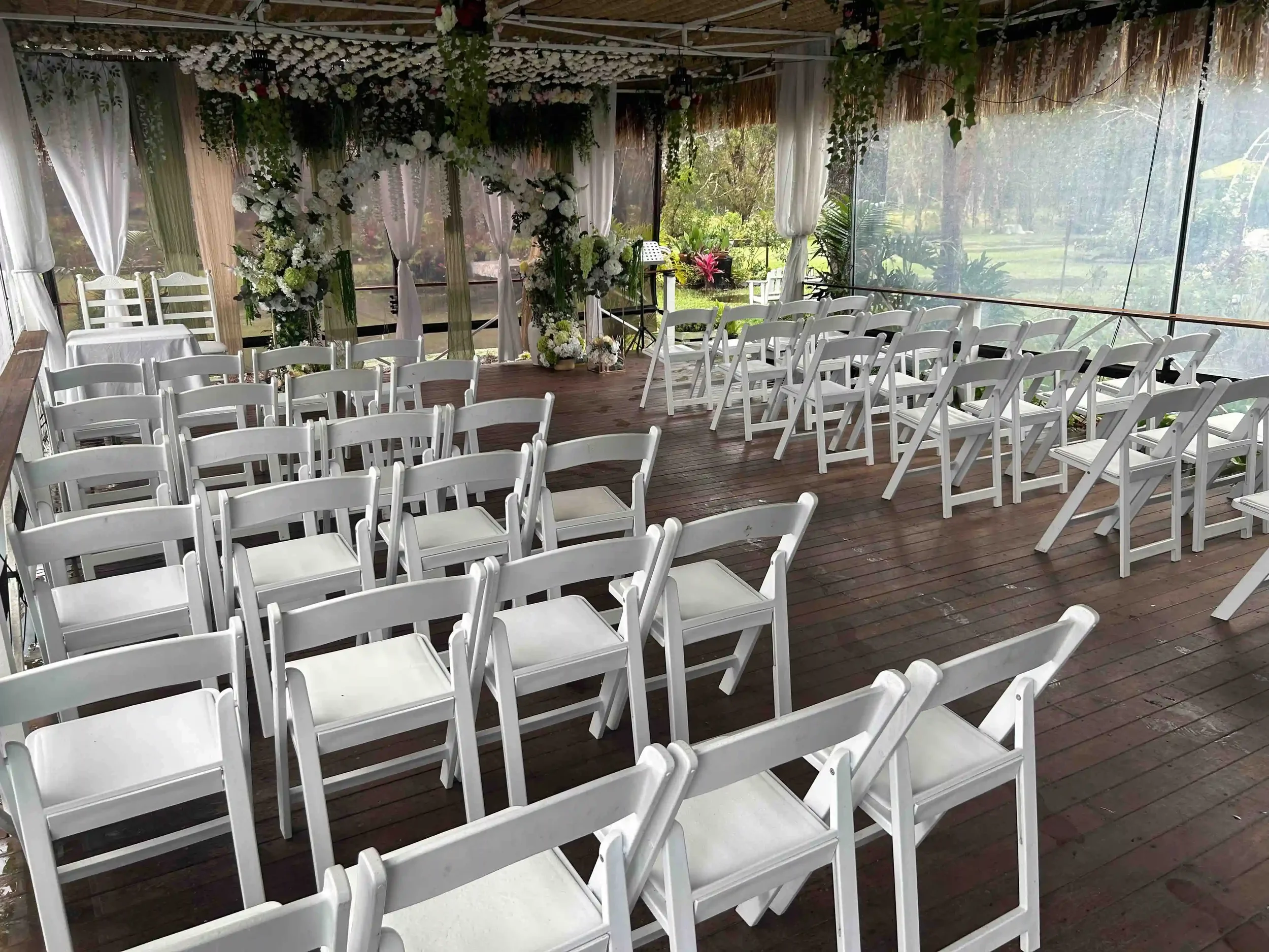 Empty white folding chairs arranged for a wedding ceremony inside a decorated outdoor pavilion with floral arrangements and curtains, overlooking a lush garden.