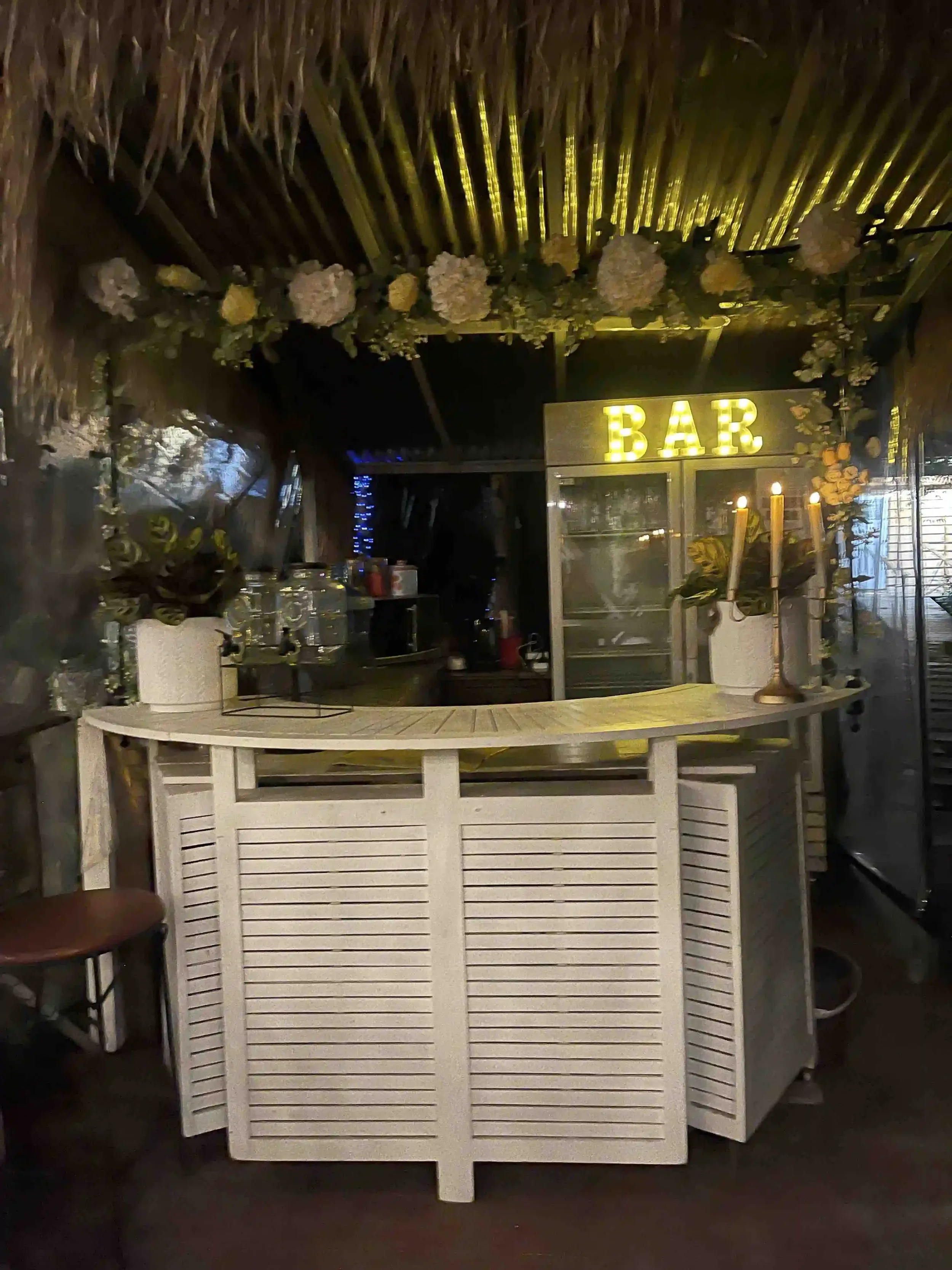 A bar counter with a white wooden foldable partition in front. In the background, there is a sign with the word 'BAR' illuminated in yellow, floral decorations on the ceiling, and some bottles and glasses on the counter.
