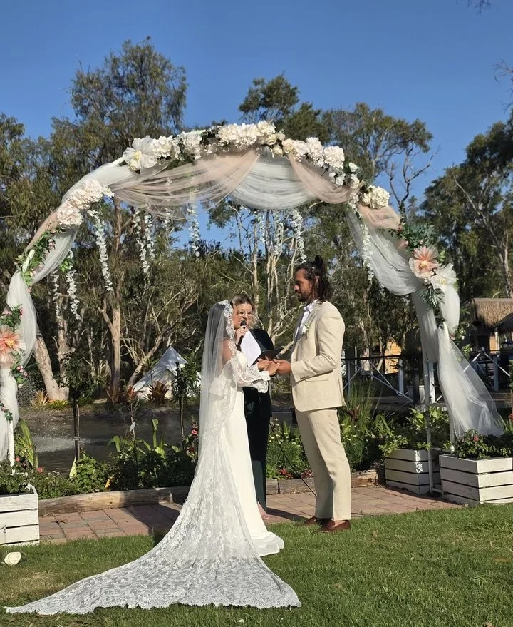 A wedding ceremony outdoors under a floral arch with a bride in a white lace dress and veil, a groom in a light beige suit. The couple is holding hands. There are trees and a blue sky in the background with flowers.