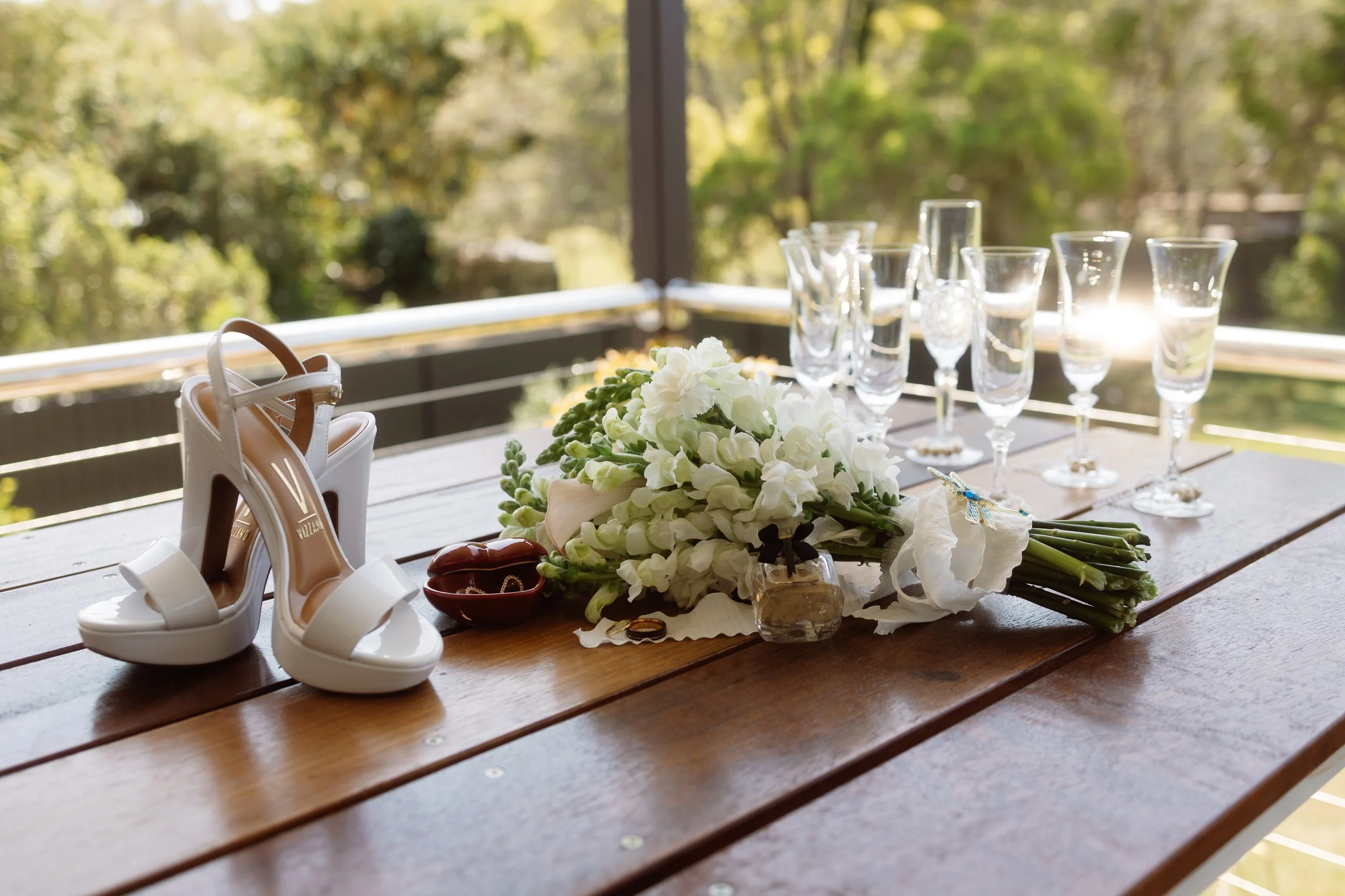 White high-heeled shoes next to a bouquet of white flowers on a wooden table with empty glassware and a small decorative item, set outdoors with sunlight and green trees in the background.